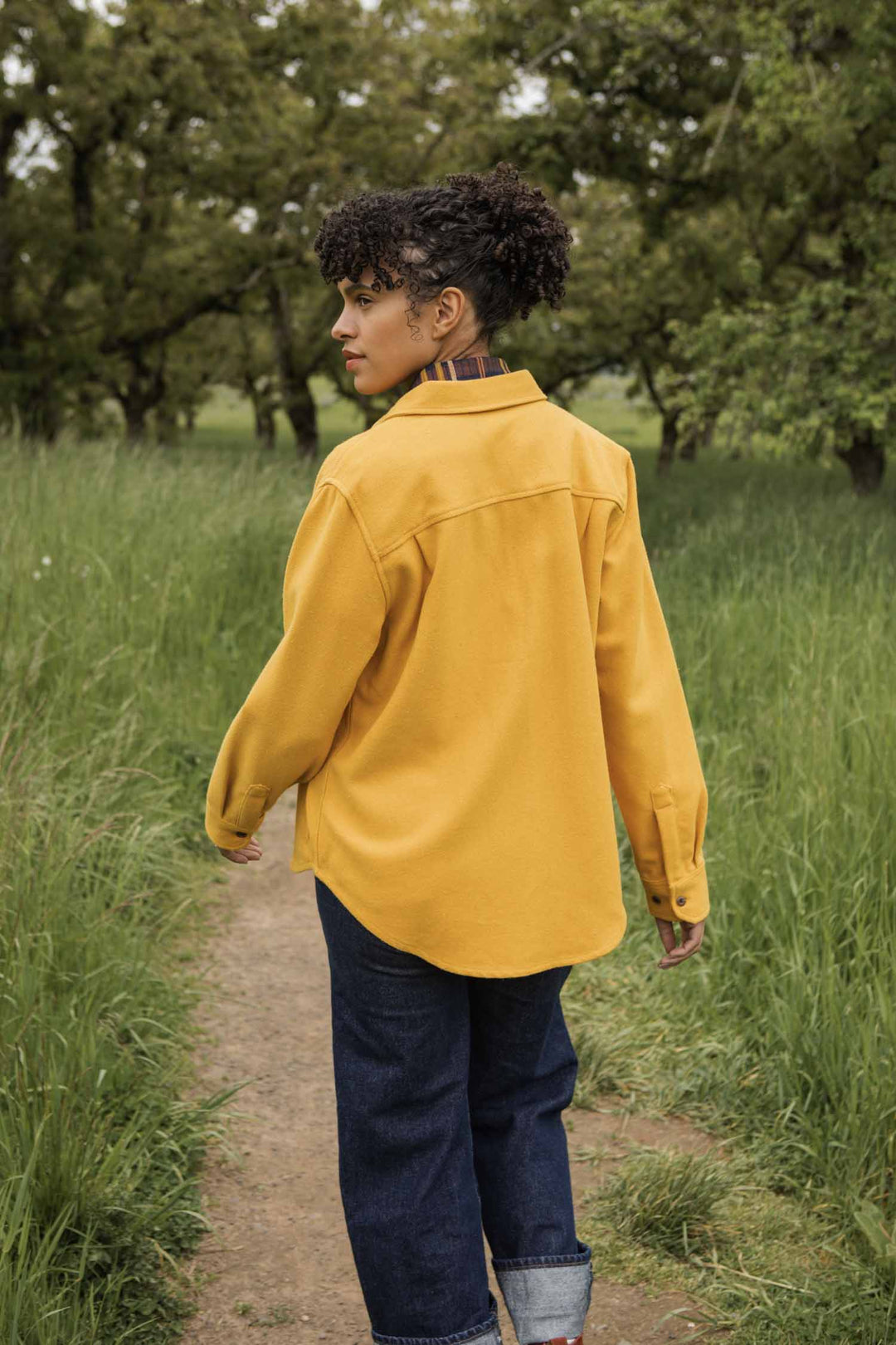 Wearing the Bridge & Burn Dawson Overshirt in Antique Gold and dark jeans, a person with curly hair walks along a dirt path through tall green grass, glancing to the side with trees in the background.
