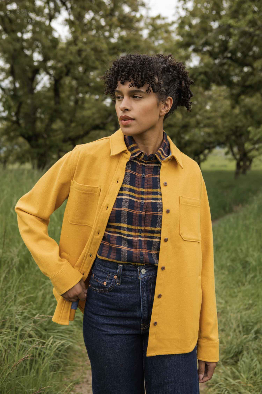 A person with curly hair stands outdoors on a path, wearing the Bridge & Burn Dawson Overshirt in Antique Gold over a recycled cotton plaid shirt and blue jeans, surrounded by trees and greenery.