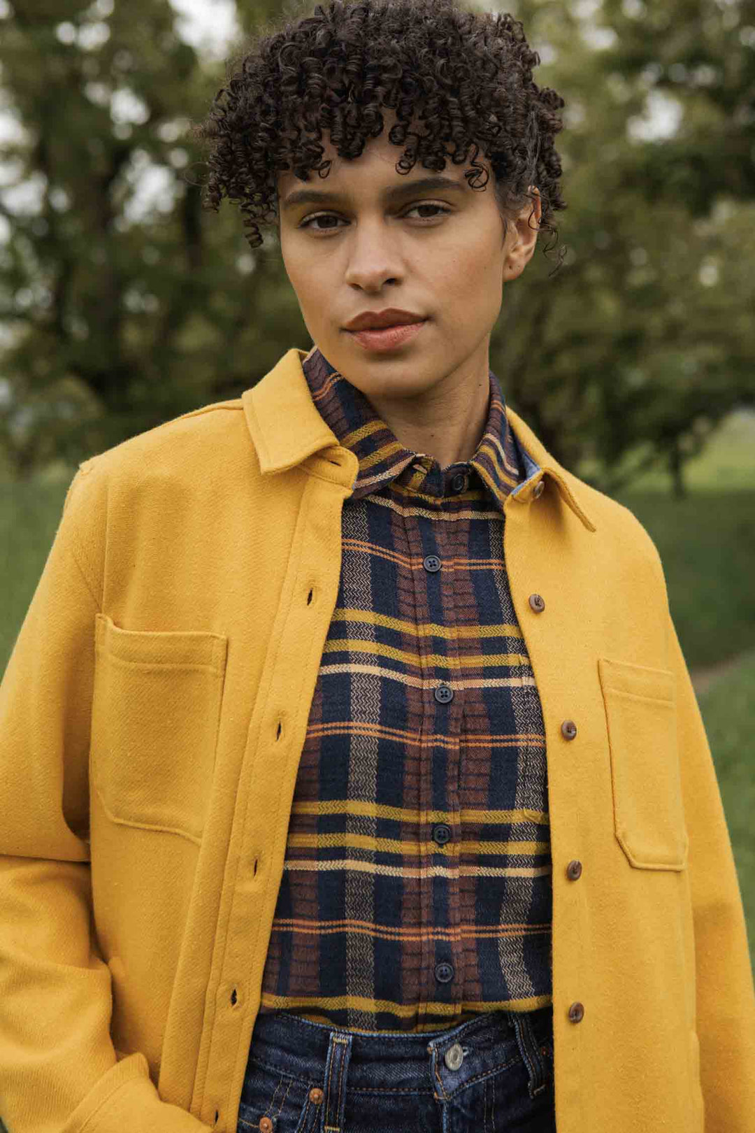 A person with short curly hair stands outdoors in front of blurred green trees, wearing Bridge & Burn's Dawson Overshirt in Antique Gold layered over a yellow jacket.