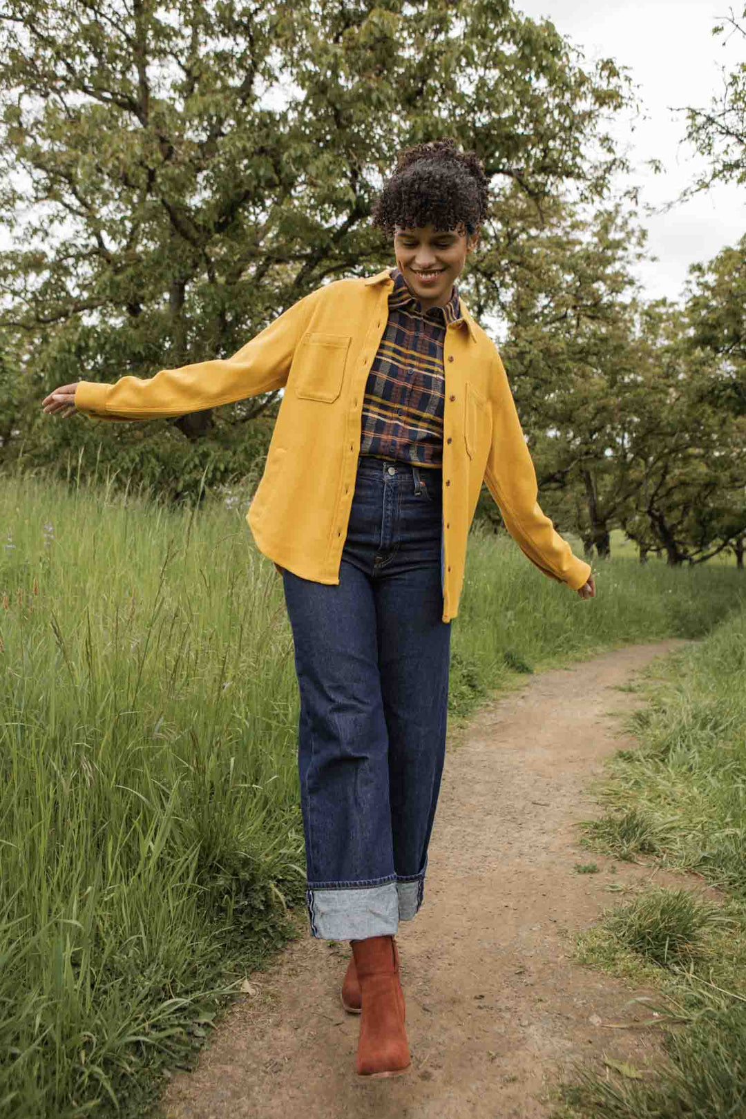 A person strolls through a grassy, tree-filled area, arms outstretched and smiling. They're wearing Bridge & Burn’s Dawson Overshirt in Antique Gold over cuffed jeans with red boots.