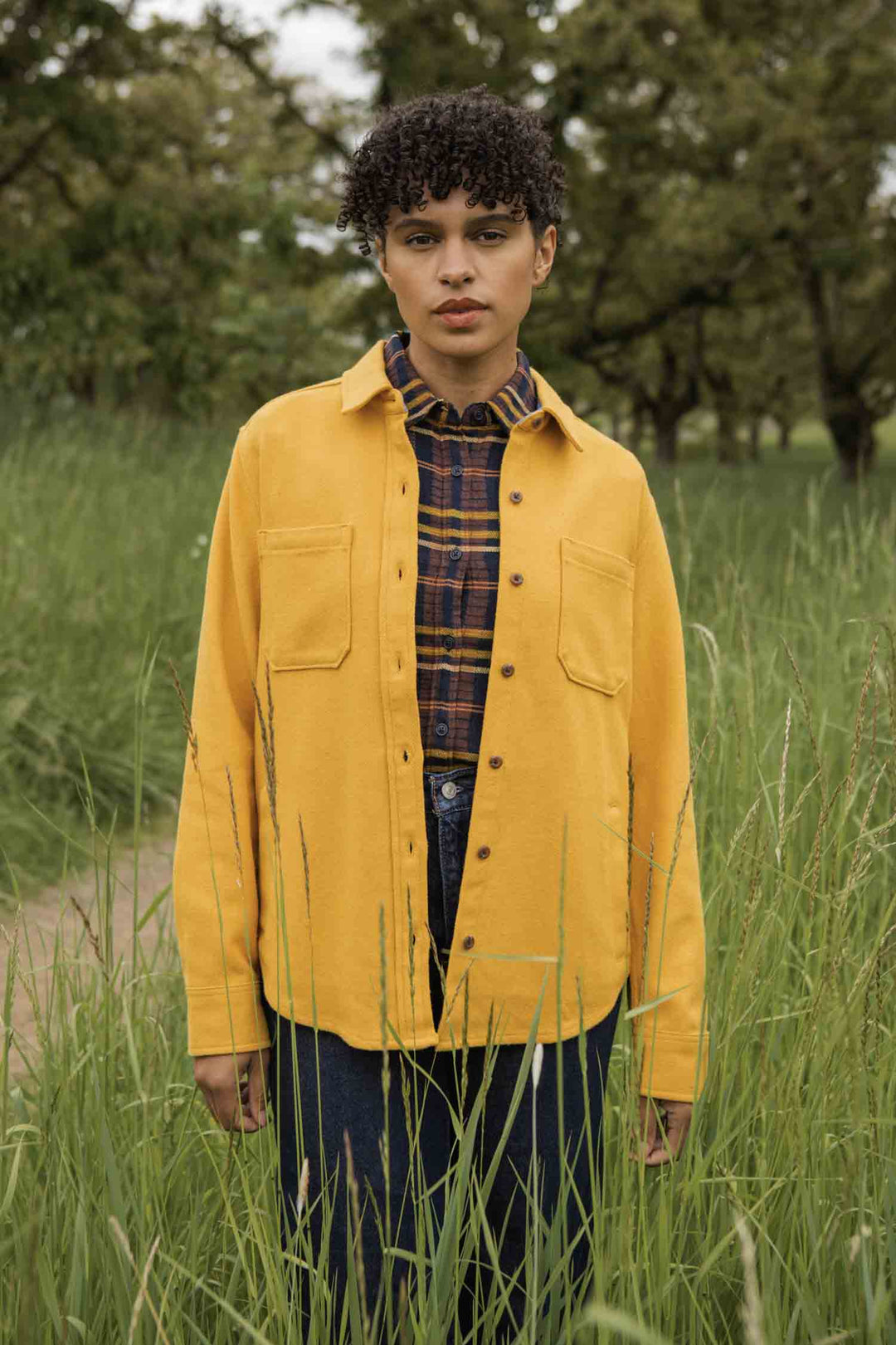 A person with short curly hair stands in tall green grass, wearing the Bridge & Burn Dawson Overshirt in Antique Gold over a plaid shirt, looking at the camera. Trees and a dirt path are visible in the background.