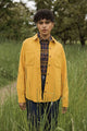 A person with short curly hair stands in tall green grass, wearing the Bridge & Burn Dawson Overshirt in Antique Gold over a plaid shirt, looking at the camera. Trees and a dirt path are visible in the background.