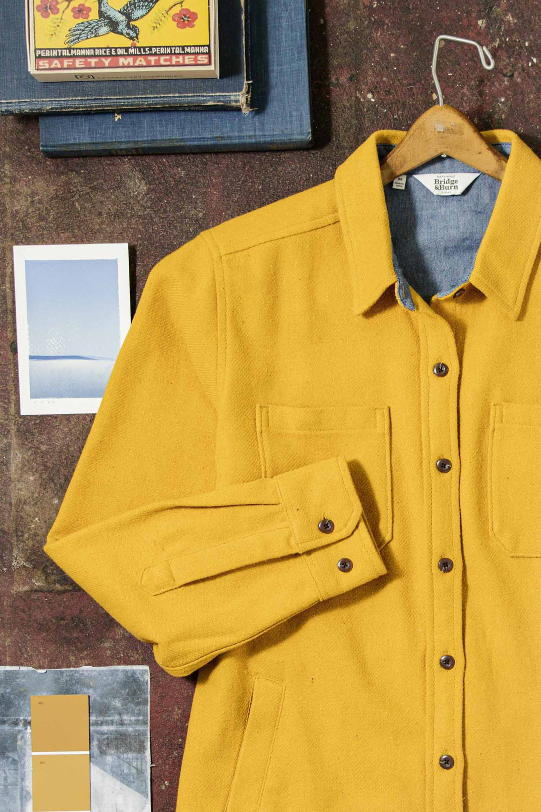 The Bridge & Burn Dawson Overshirt in Antique Gold rests on a brown surface beside a blue book, snowy landscape postcard, paint swatches, and a box with cartoon characters.