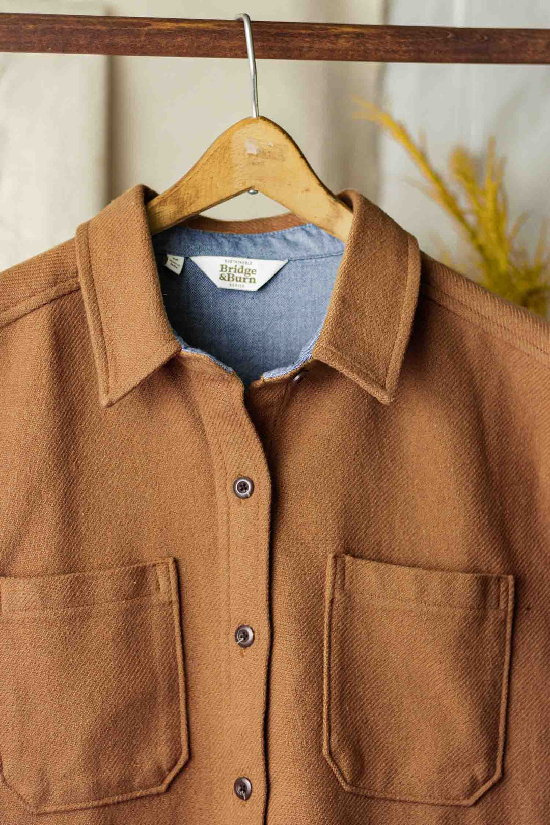 The Dawson Overshirt in camel by Bridge & Burn, featuring two chest pockets, hangs on a wooden hanger. The visible brand tag reflects their sustainable values, while a blurred yellow plant appears in the background.