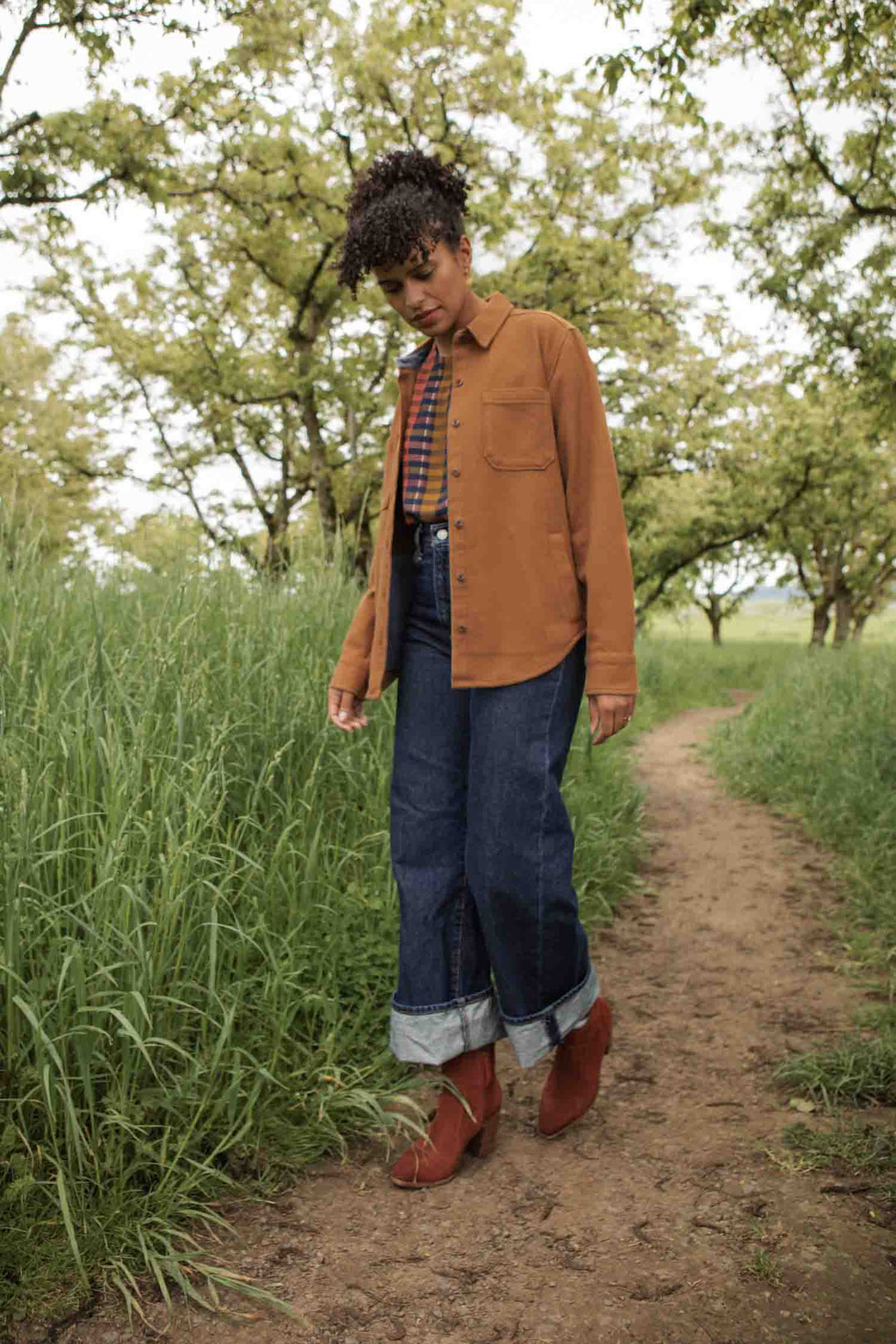 A person walks on a dirt path in tall grass and trees, wearing cuffed jeans, brown boots, and the Bridge & Burn Dawson Overshirt in Camel, looking down thoughtfully.