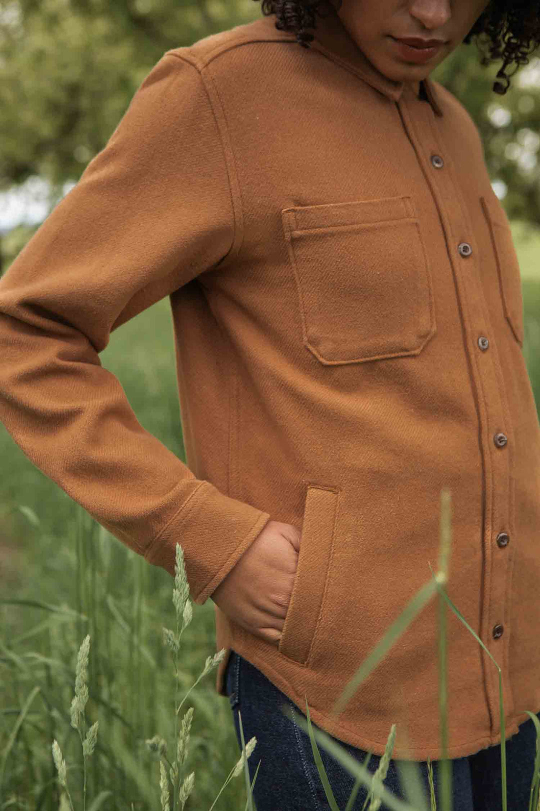 Outdoors in tall grass, a person wears the Bridge & Burn Dawson Overshirt in Camel, featuring chest and side pockets. One hand is tucked in; their face is partially out of frame.