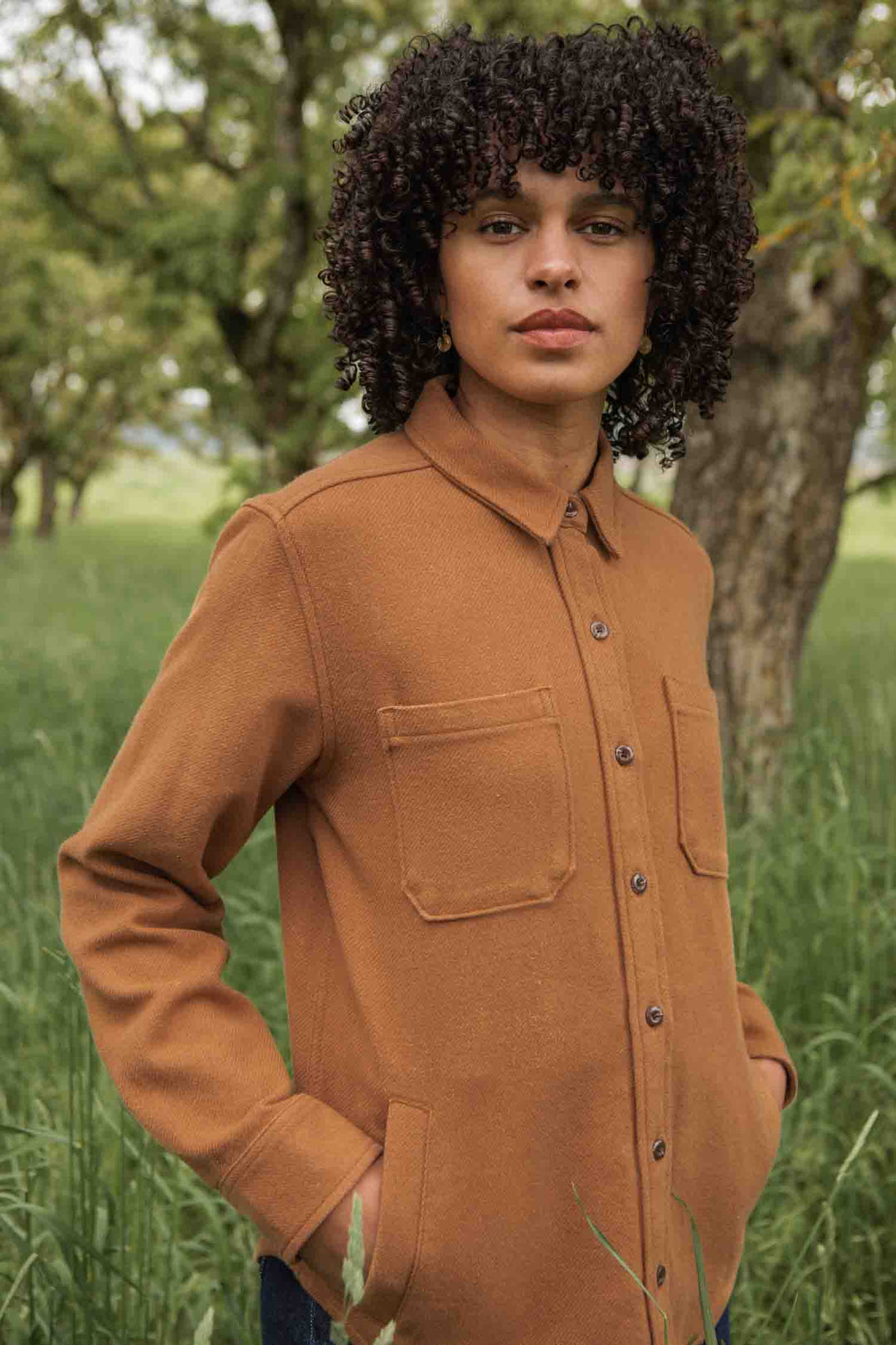 A person with curly hair wears the Bridge & Burn Dawson Overshirt in Camel, made from recycled cotton, while standing outdoors on a grassy area with trees behind them and looking at the camera.