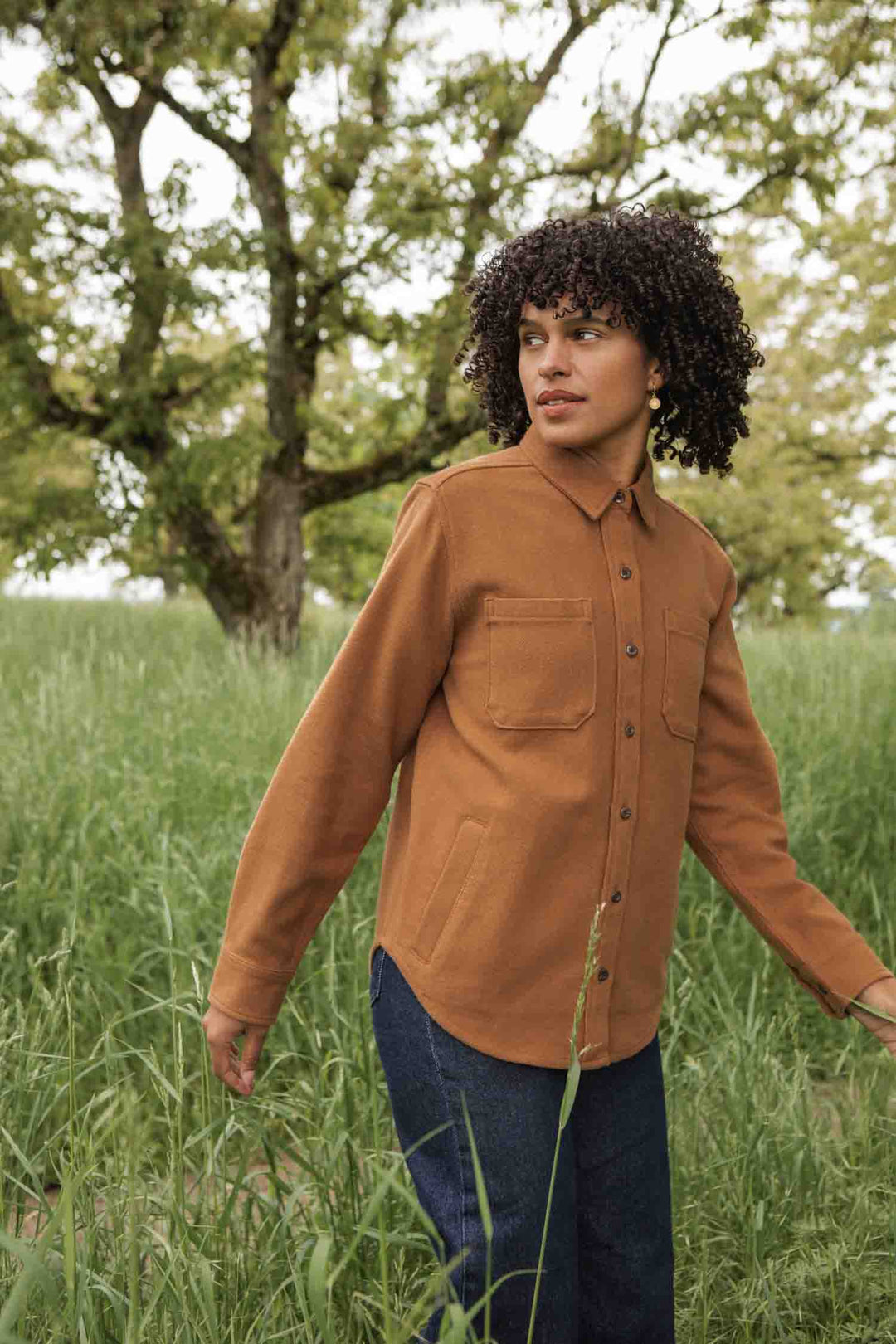 A person with curly hair wears the Bridge & Burn Dawson Overshirt in Camel and blue jeans, standing in tall grass and looking to the side, with trees and greenery in the background.
