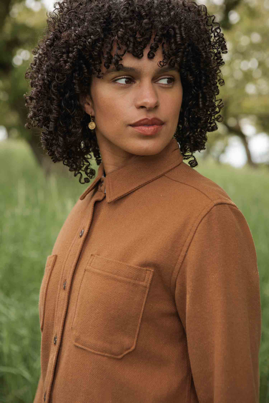 A person with curly dark hair stands outside in green grass and blurred trees, wearing the Bridge & Burn Dawson Overshirt in Camel.
