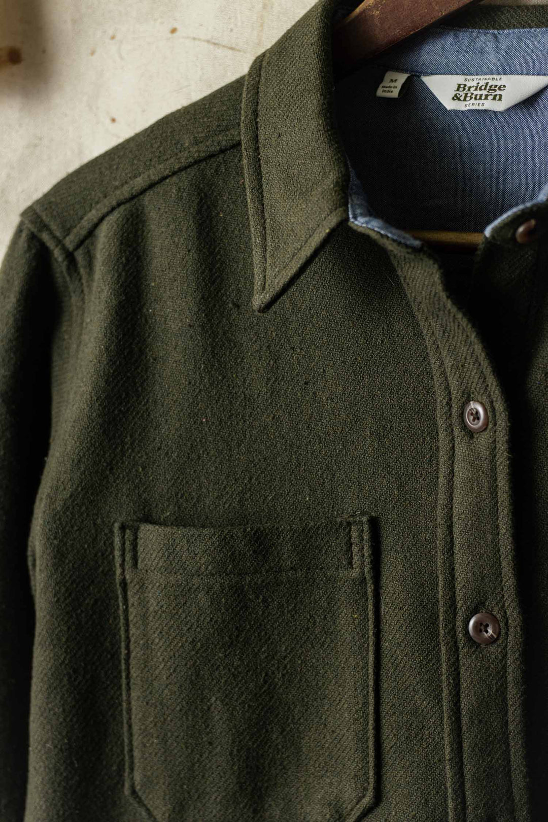 A close-up of the Bridge & Burn Dawson Overshirt in Dark Sage, featuring a collar and chest pocket, hanging on a wooden hanger with the brand label partially visible.