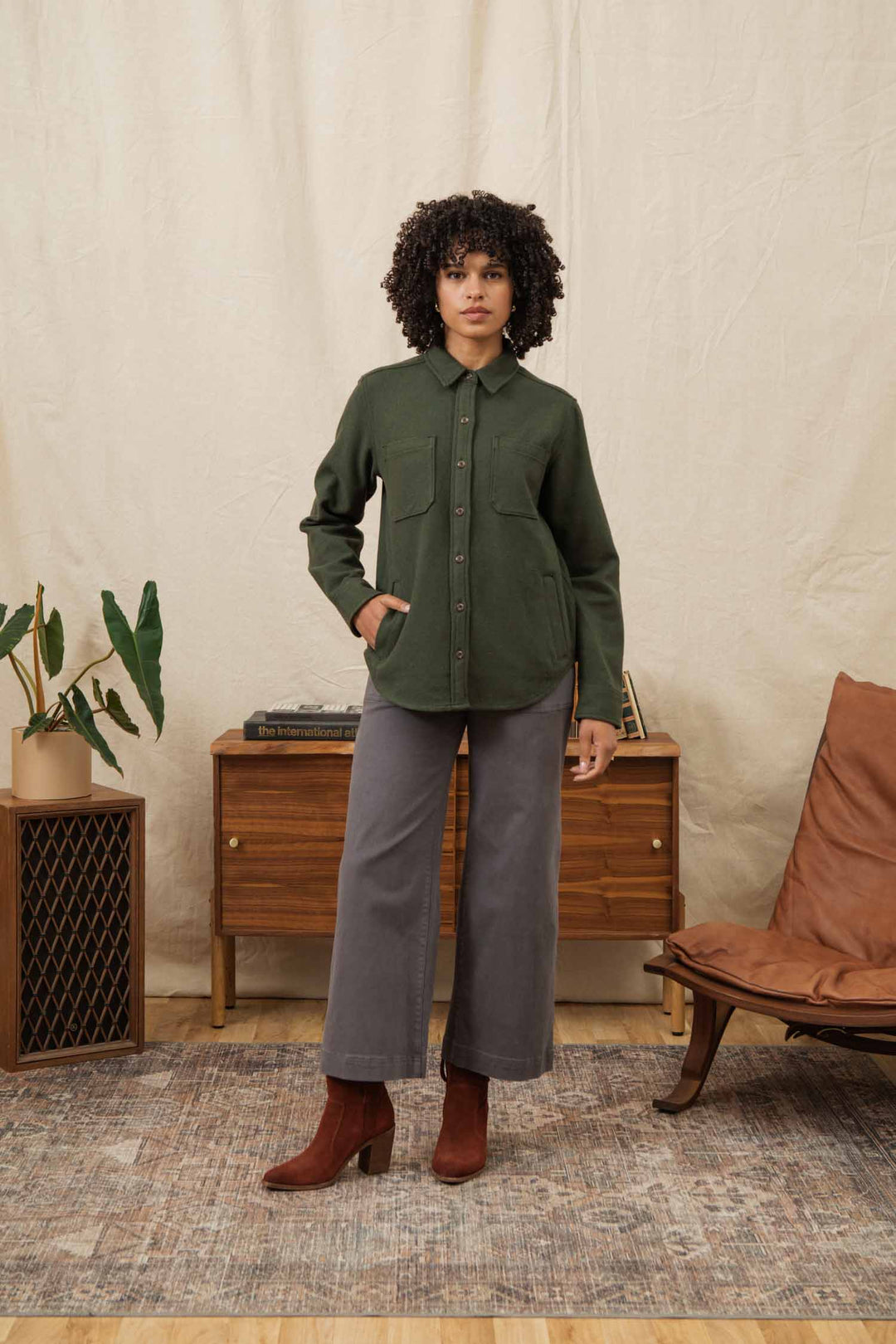 A woman with curly hair stands indoors on a rug, wearing the Bridge & Burn Dawson Overshirt in Dark Sage, gray wide-leg pants, and brown heeled boots. She poses by a wooden cabinet and tan chair with one hand in her pocket.