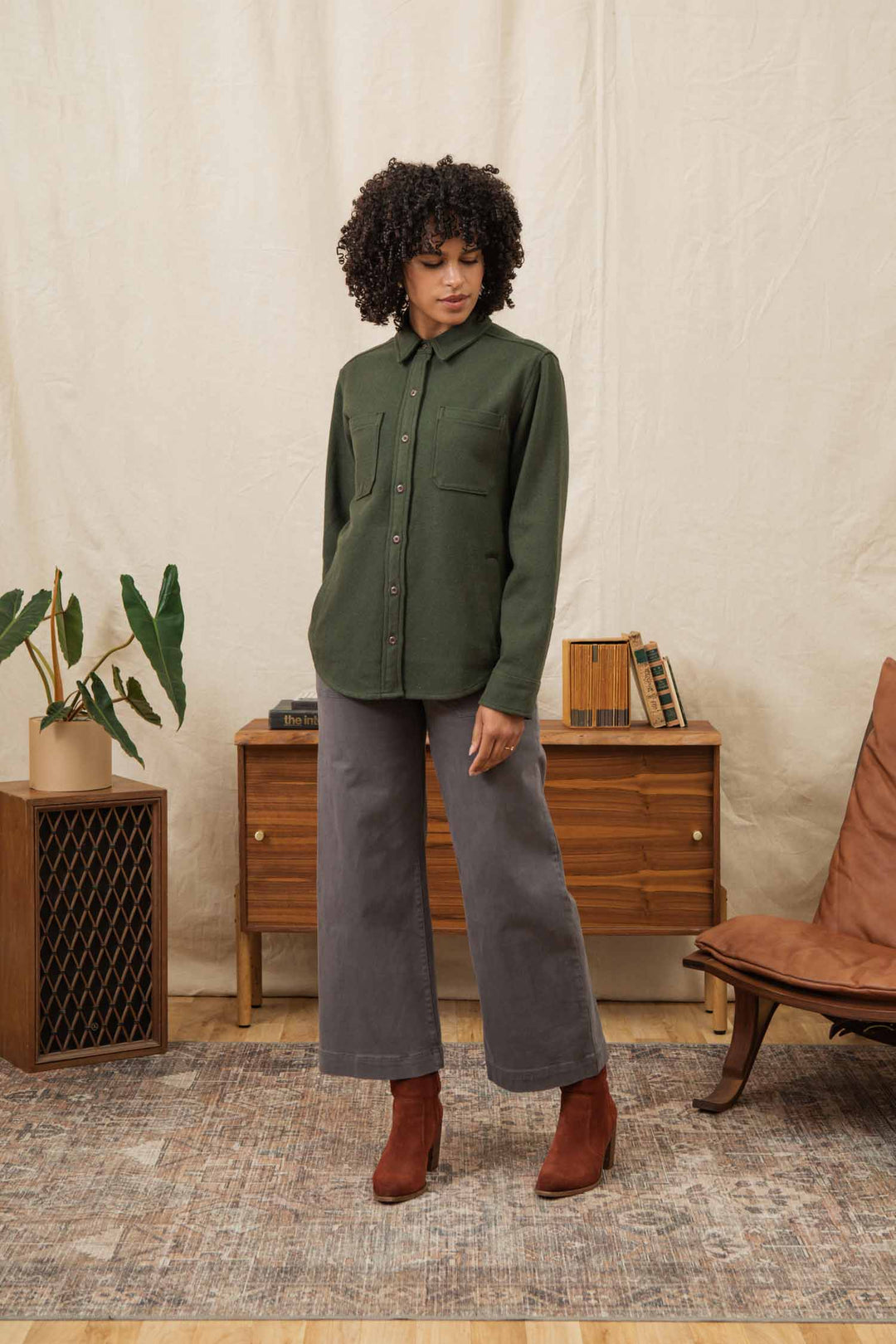 A woman with curly hair stands indoors on a patterned rug, wearing the Bridge & Burn Dawson Overshirt in Dark Sage, gray wide-leg pants, and brown ankle boots. Behind her are a wooden cabinet, plant, books, and a brown chair.