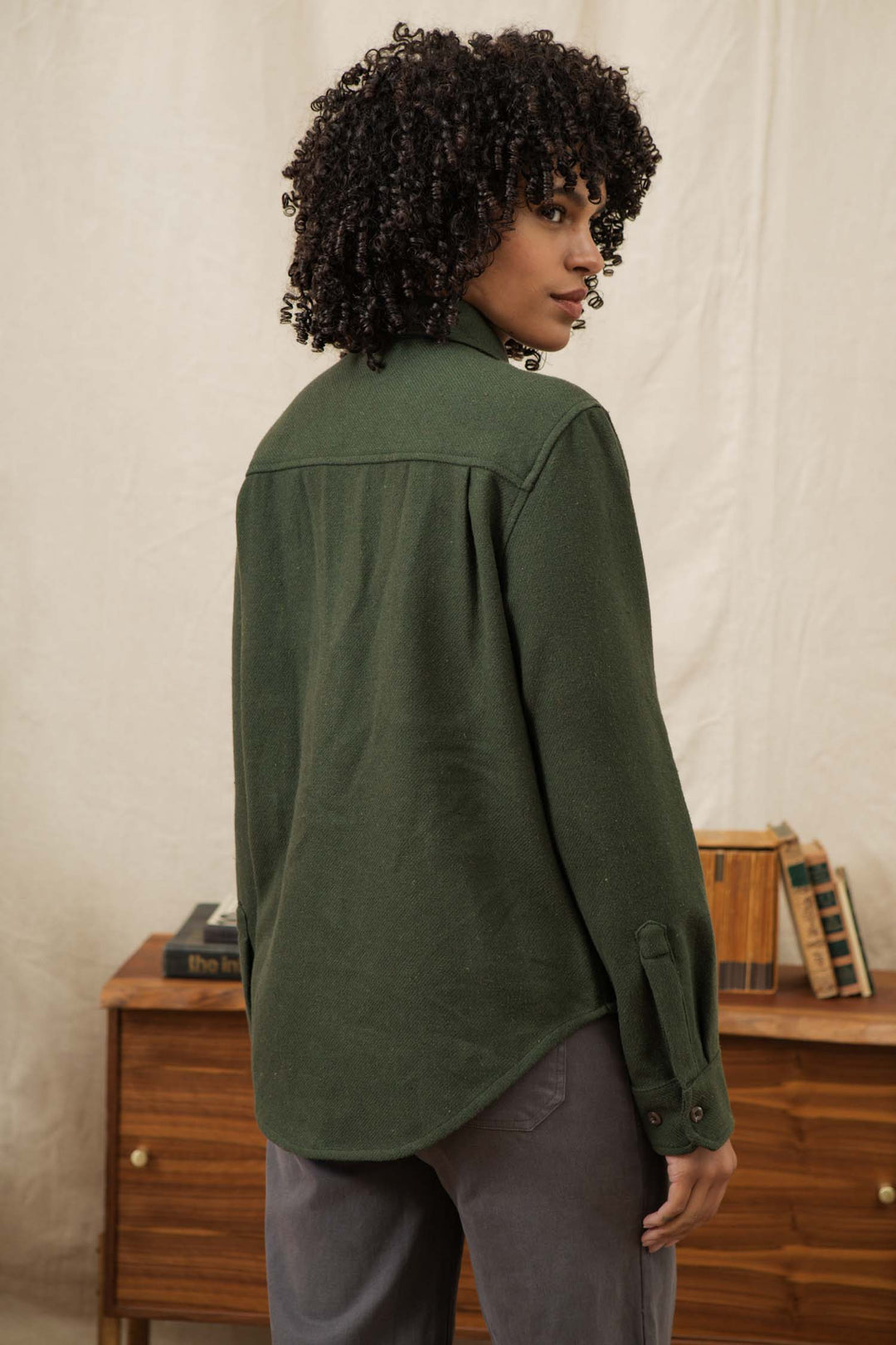 A person with curly hair stands indoors, looking over their shoulder. They are wearing the Bridge & Burn Dawson Overshirt in Dark Sage and gray pants. A wooden table with books is visible in the background.