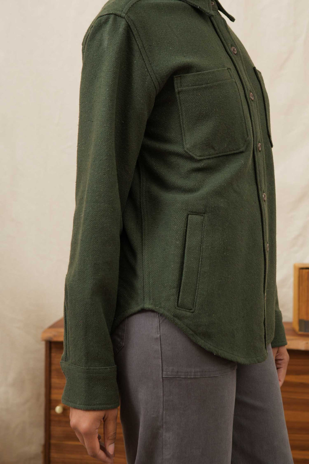 A person stands sideways in the Bridge & Burn Dawson Overshirt in Dark Sage, crafted from recycled cotton with chest and side pockets, paired with gray pants. A wooden cabinet and beige wall are in the background.
