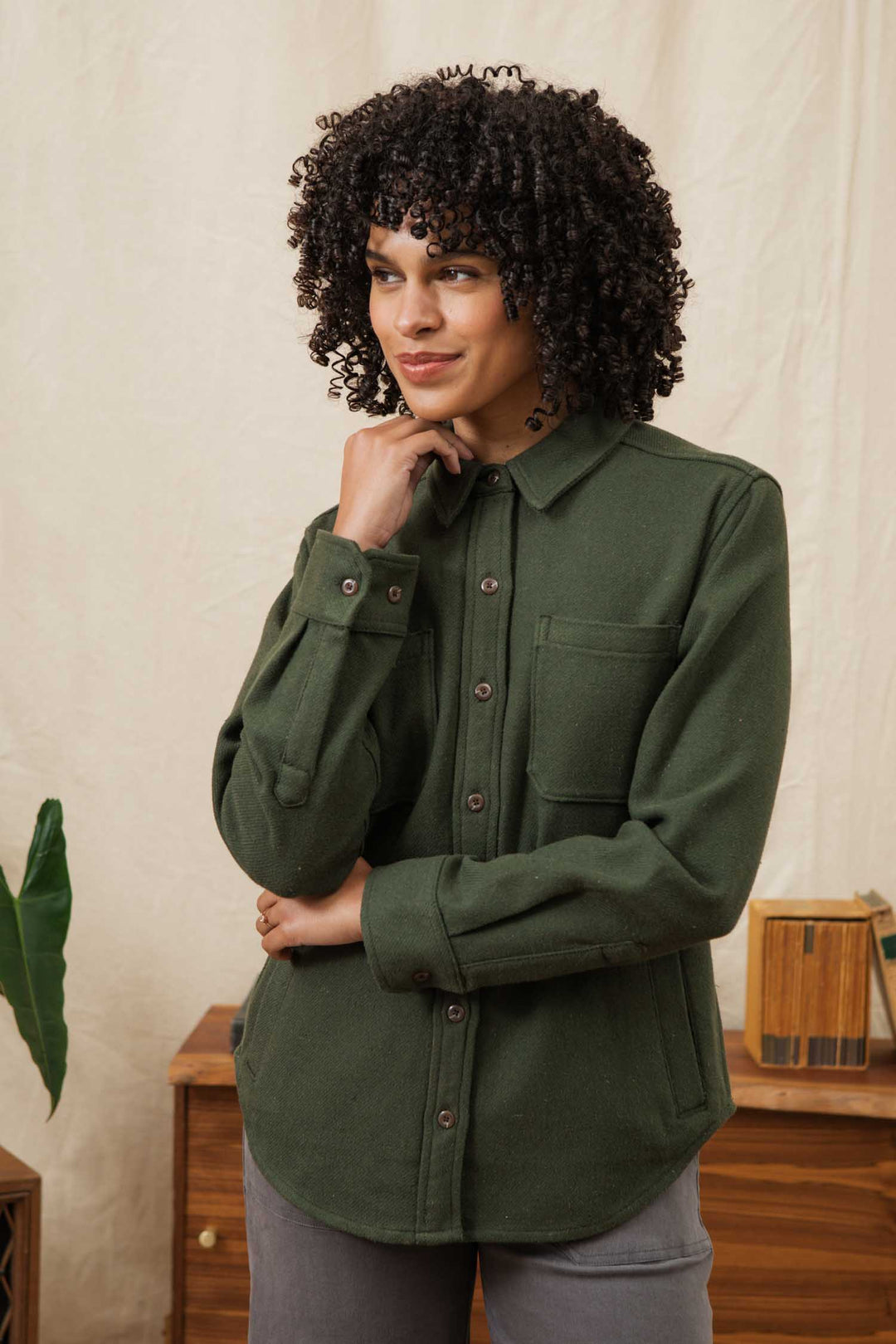 A person with curly hair wears the Bridge & Burn Dawson Overshirt in Dark Sage and gray pants, standing indoors by a wooden table and cream backdrop, one hand on their chin and the other arm crossed.