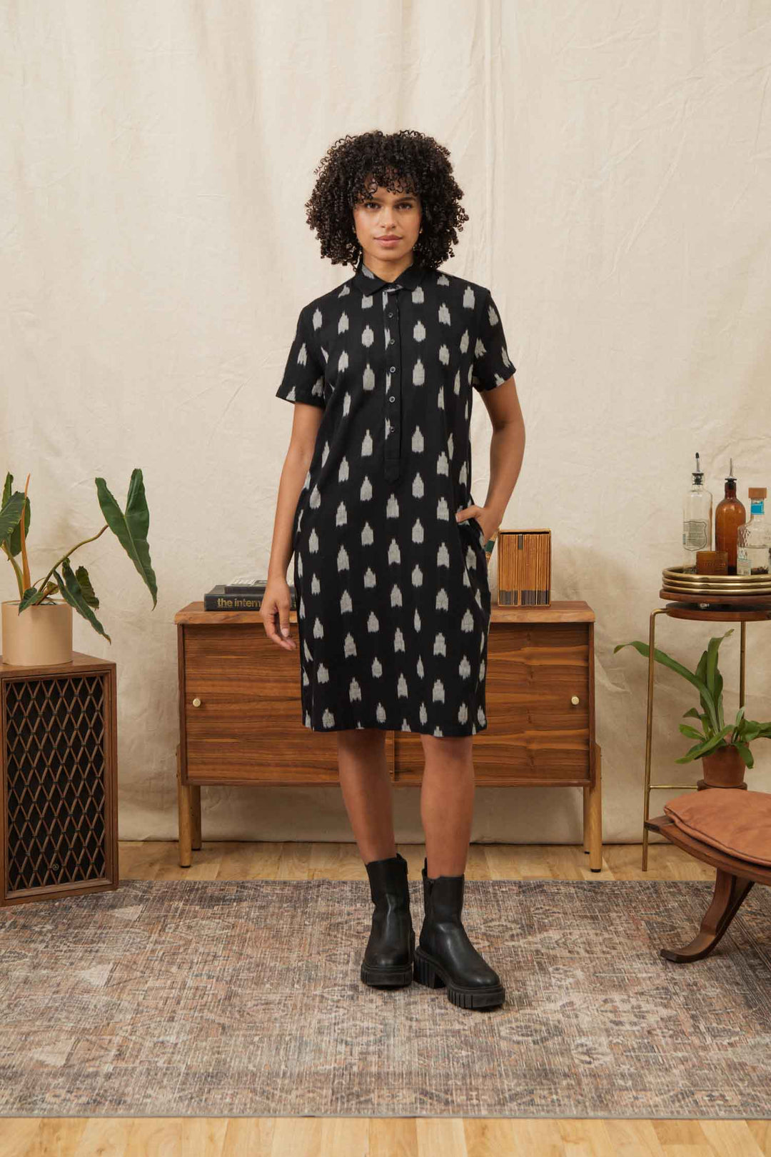 A person with curly hair wears the Bridge & Burn Delia Shirt Dress in Eclipse Ikat and black boots, standing in a cozy room with mid-century furniture, plants, and a rug on a wood floor.