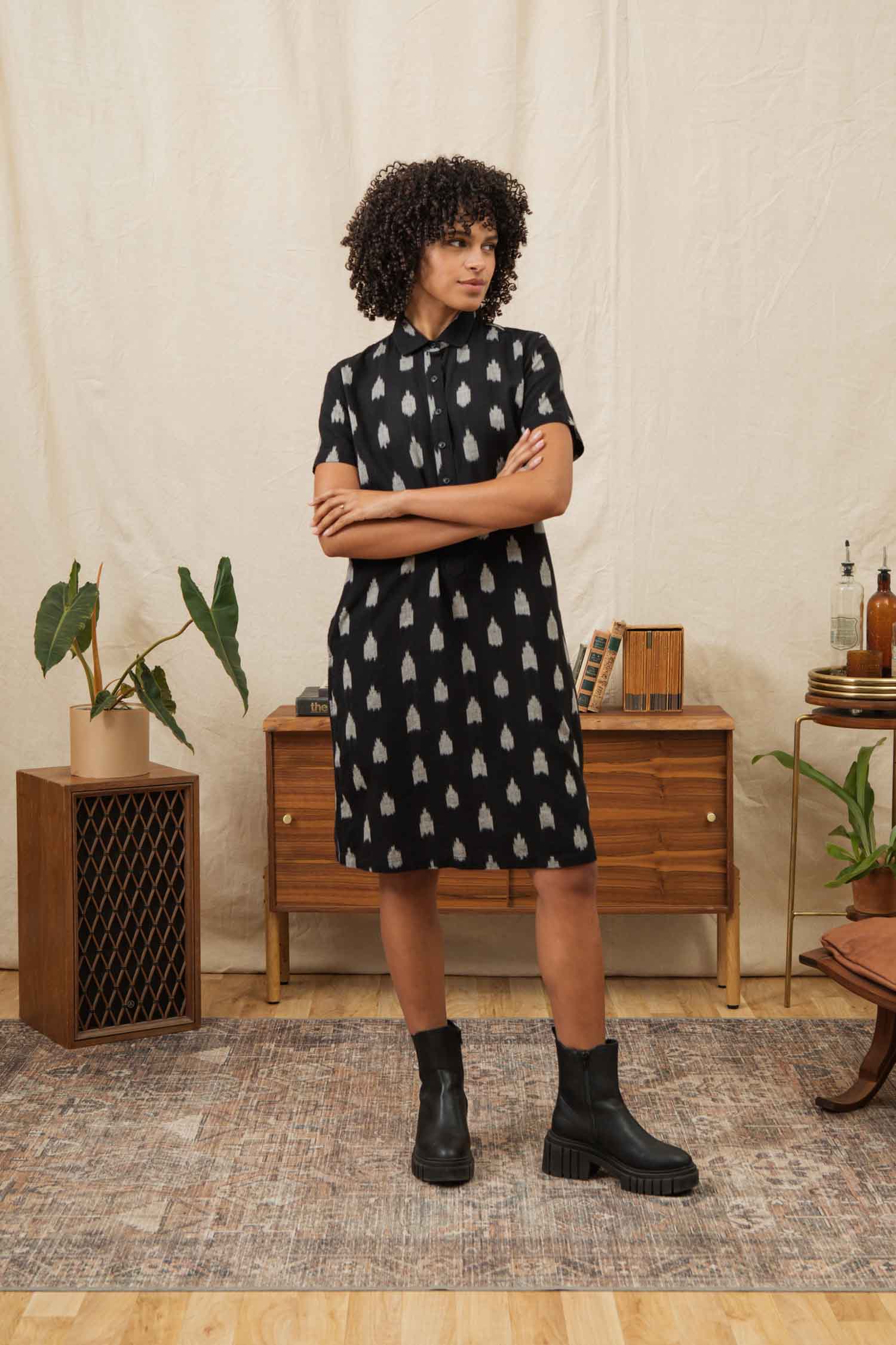 A woman with curly hair stands with arms crossed, wearing the Bridge & Burn Delia Shirt Dress / Eclipse Ikat and black boots in a cozy room decorated with wooden furniture, plants, and a rug.