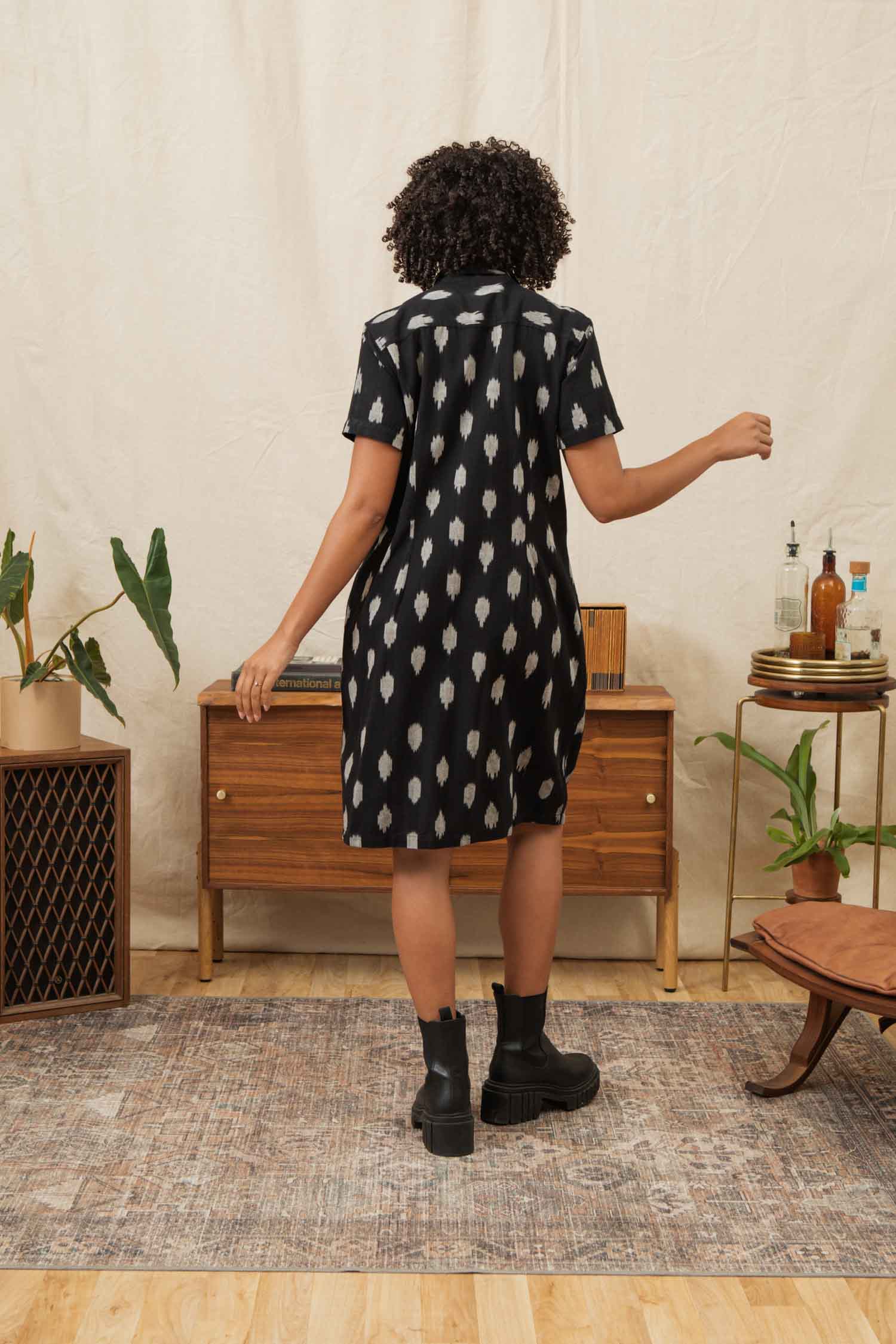 A person with curly hair, wearing the Bridge & Burn Delia Shirt Dress in Eclipse Ikat and black boots, stands with their back to the camera in a plant-filled living room with a wooden cabinet and decor.