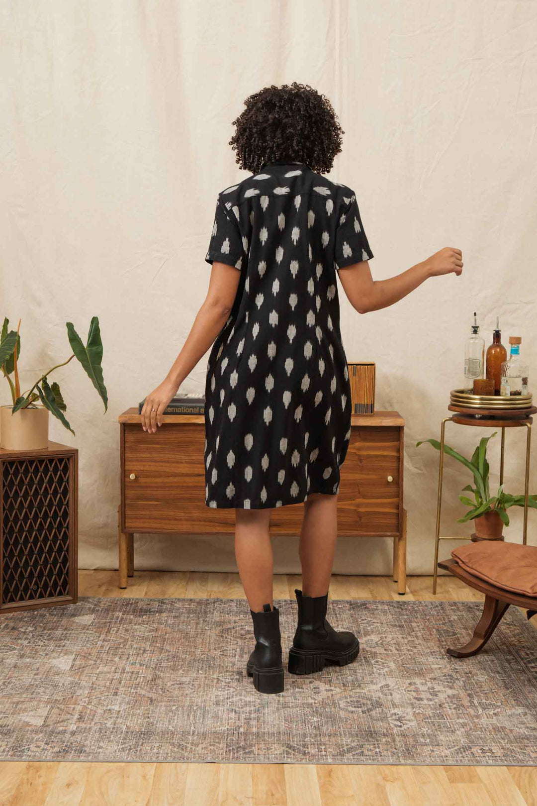 A person with curly hair, wearing the Bridge & Burn Delia Shirt Dress in Eclipse Ikat and black boots, stands with their back to the camera in a plant-filled living room with a wooden cabinet and decor.