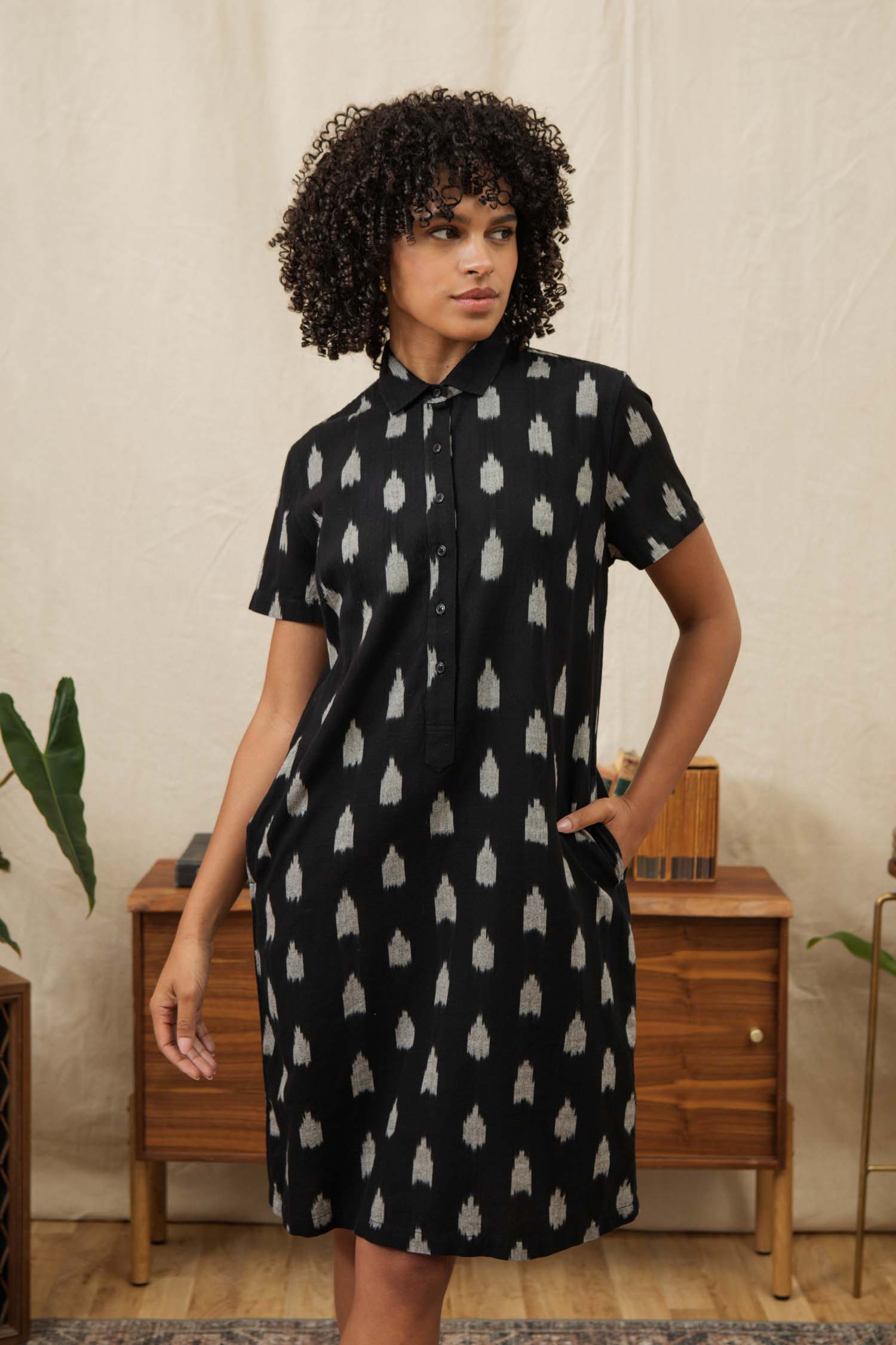 A woman with curly hair wears the Bridge & Burn Delia Shirt Dress in Eclipse Ikat, standing indoors by a wooden cabinet and plants, looking to the side with one hand in her pocket.