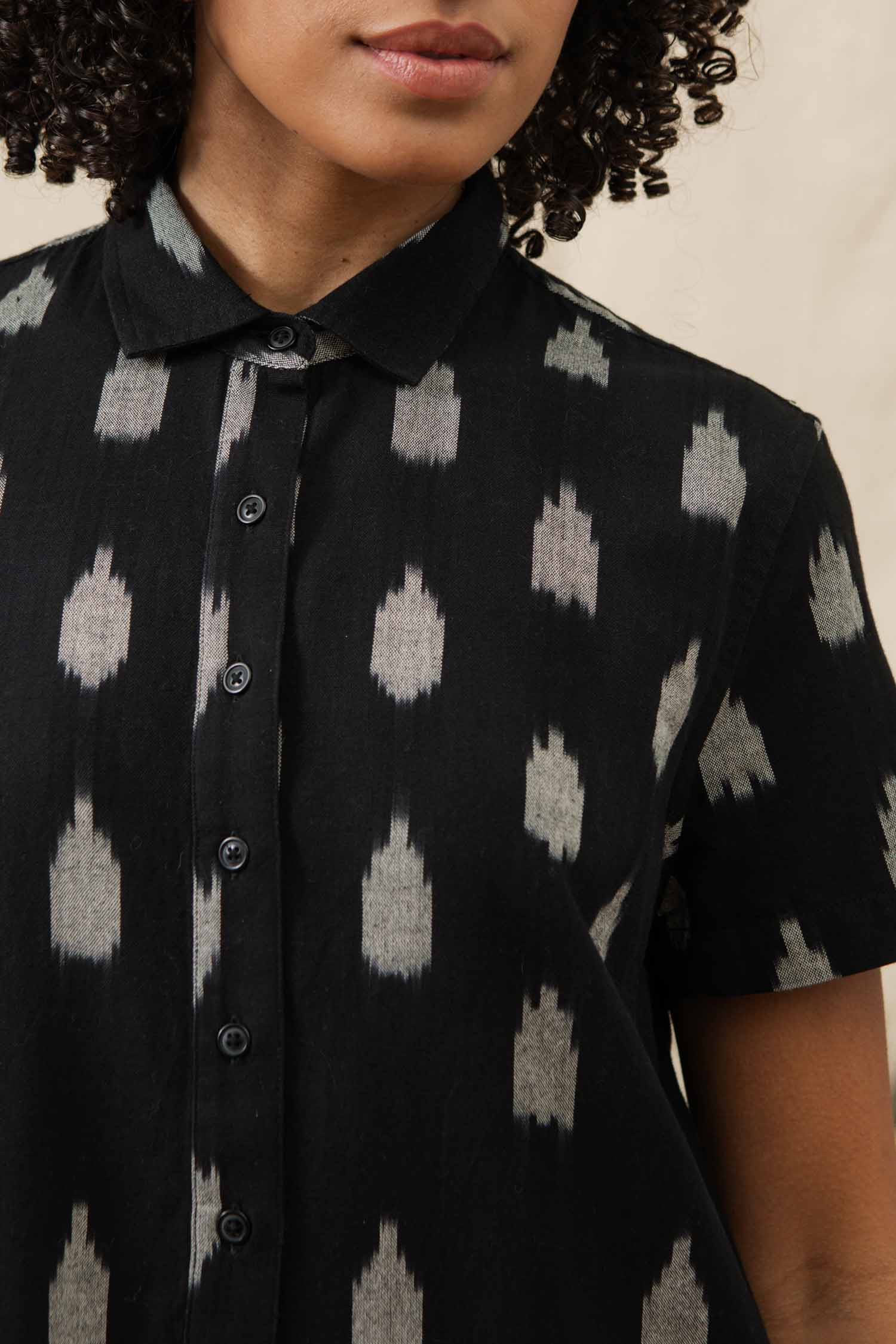 A person with curly hair wears the Bridge & Burn Delia Shirt Dress in Eclipse Ikat, a short-sleeved black button-up made from organic cotton with a light gray geometric pattern. Only the lower half of the face and upper torso are visible.