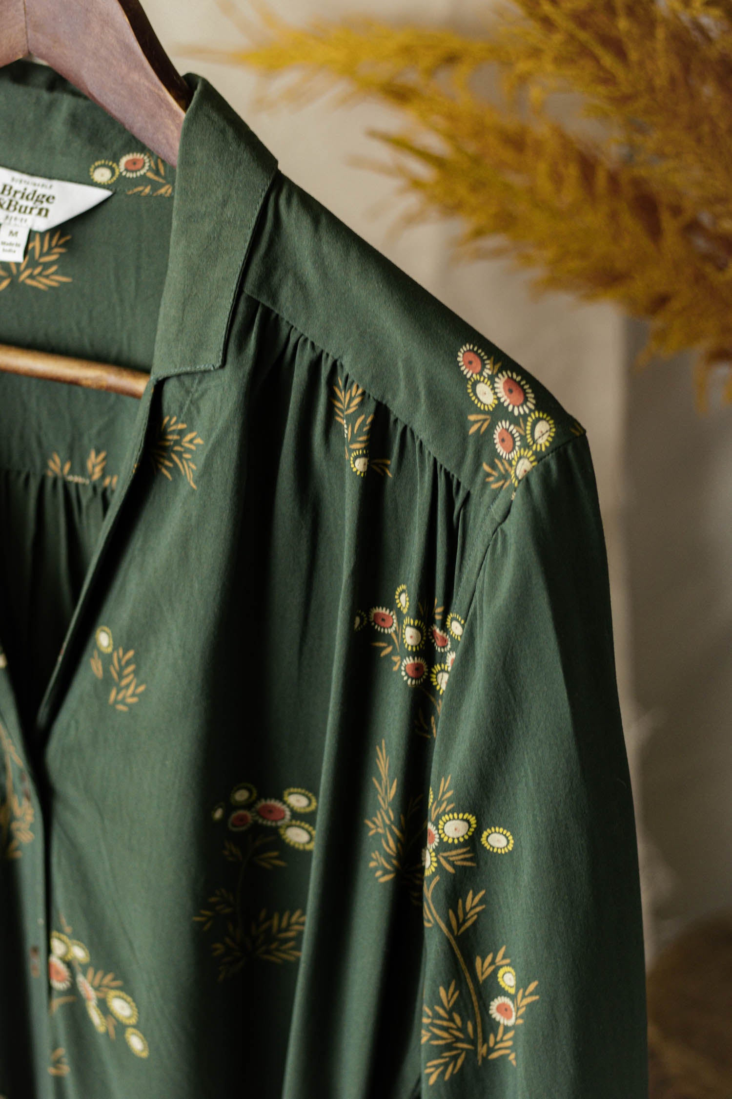 A close-up of the Bridge & Burn Emery Shirt Dress in Woodland Bloom, a green v-neck shirtdress with yellow and red floral patterns, hangs on a wooden hanger against a beige background with dried grass.