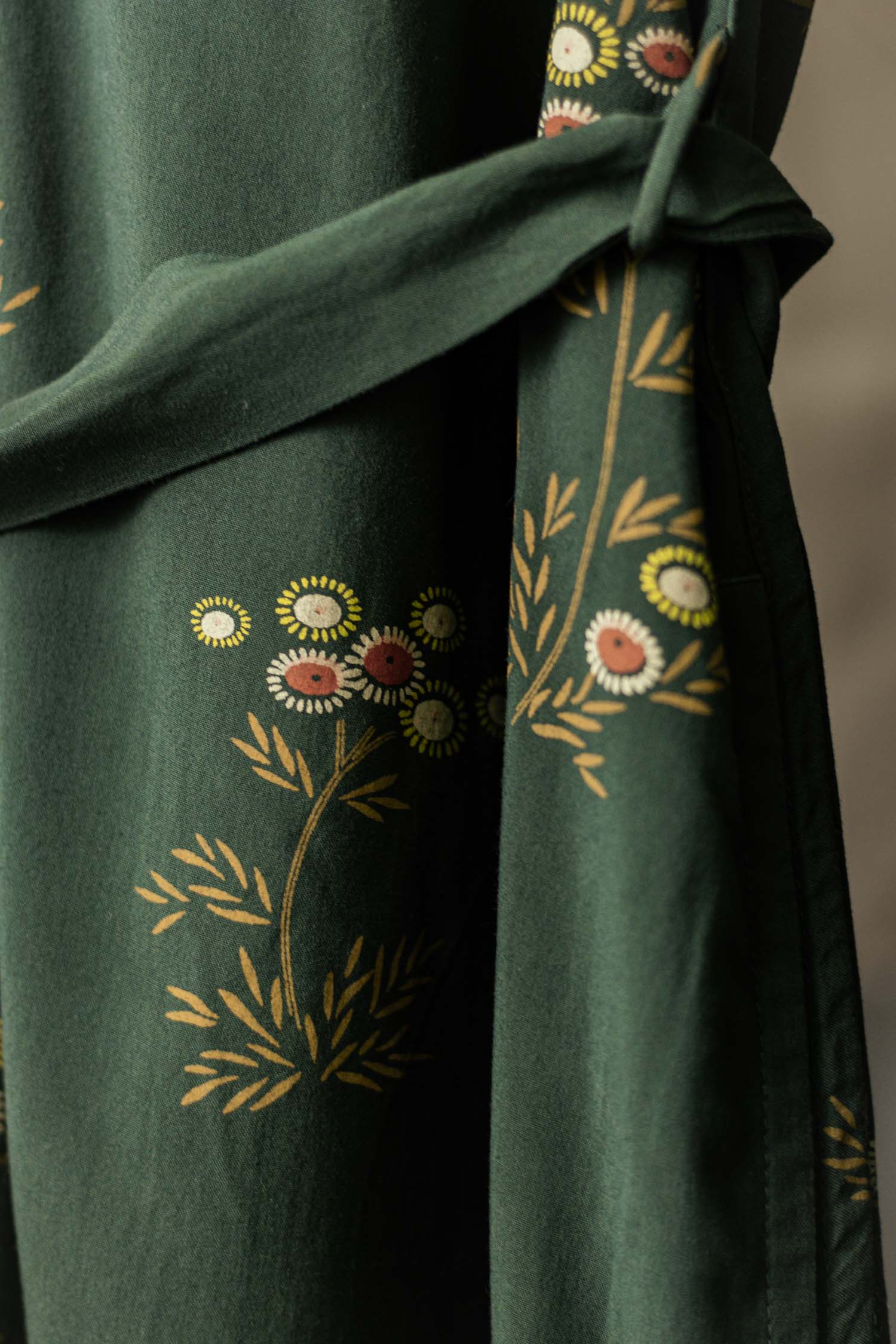 The Bridge & Burn Emery Shirt Dress in Woodland Bloom is a dark green, eco-friendly v-neck dress with a floral print in yellow, white, and red, featuring leafy branches and small daisies. It includes a belt or sash for added charm.
