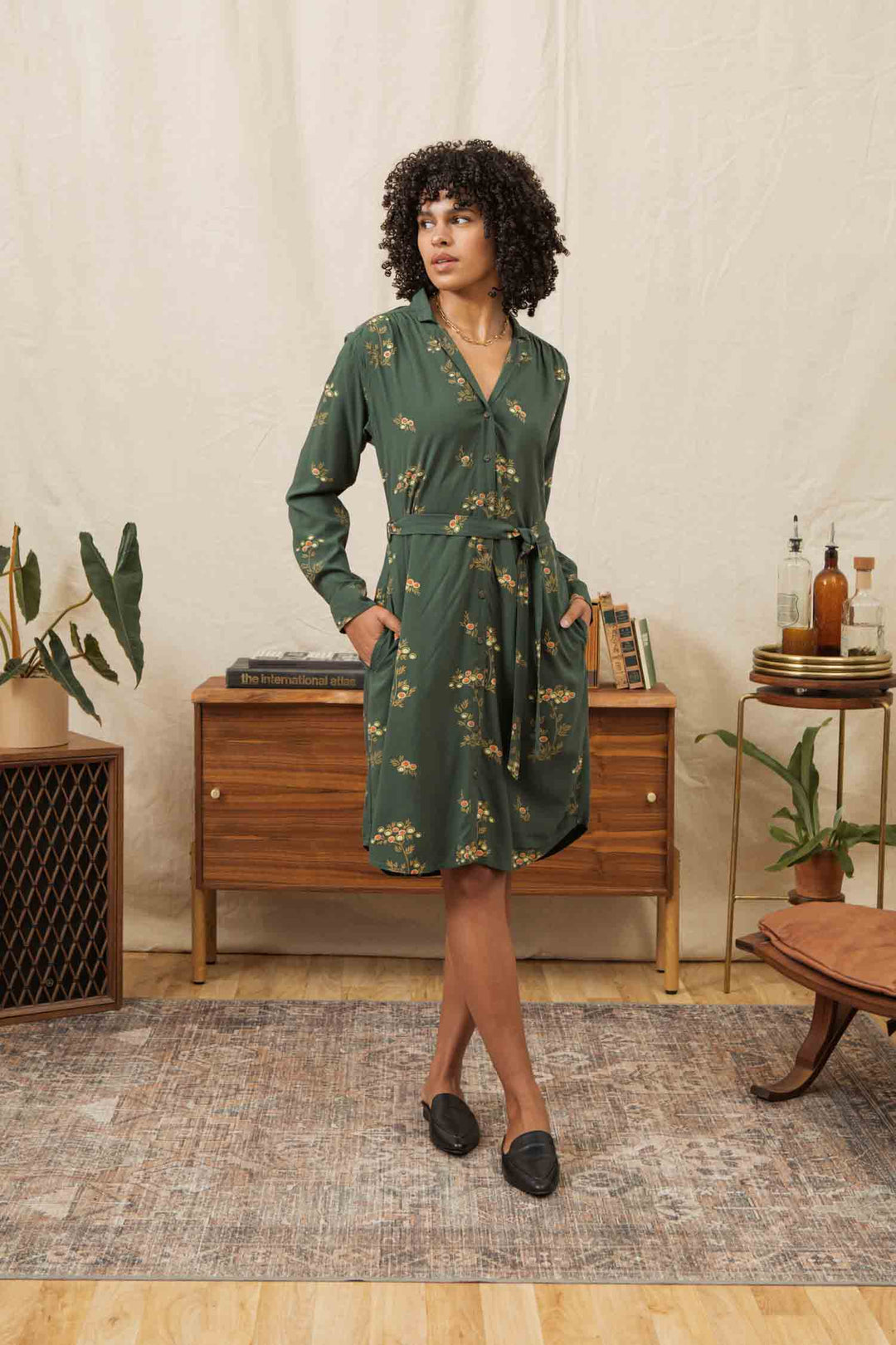 A woman with curly hair wears the Bridge & Burn Emery Shirt Dress in Woodland Bloom and black flats, standing in a stylish living room with mid-century furniture, houseplants, and a decorative rug.