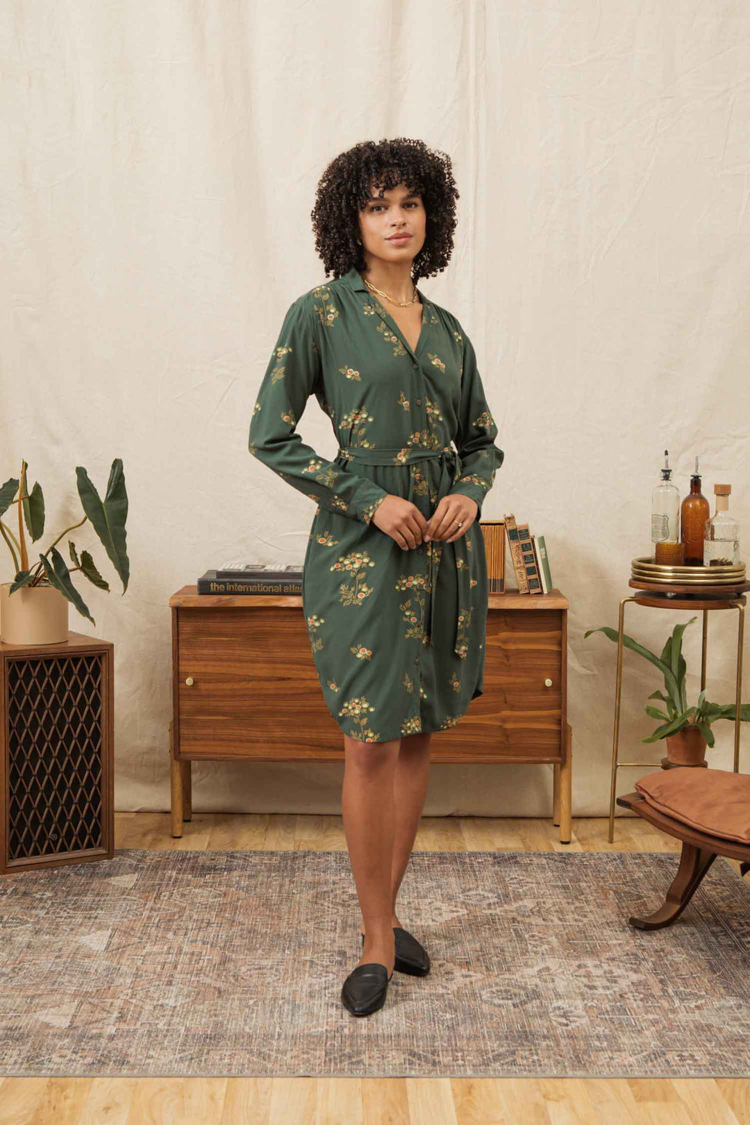 A woman with curly hair wears the Bridge & Burn Emery Shirt Dress in Woodland Bloom and black shoes, standing in a living room featuring a wooden dresser, books, plants, and a bar cart against a neutral fabric backdrop.