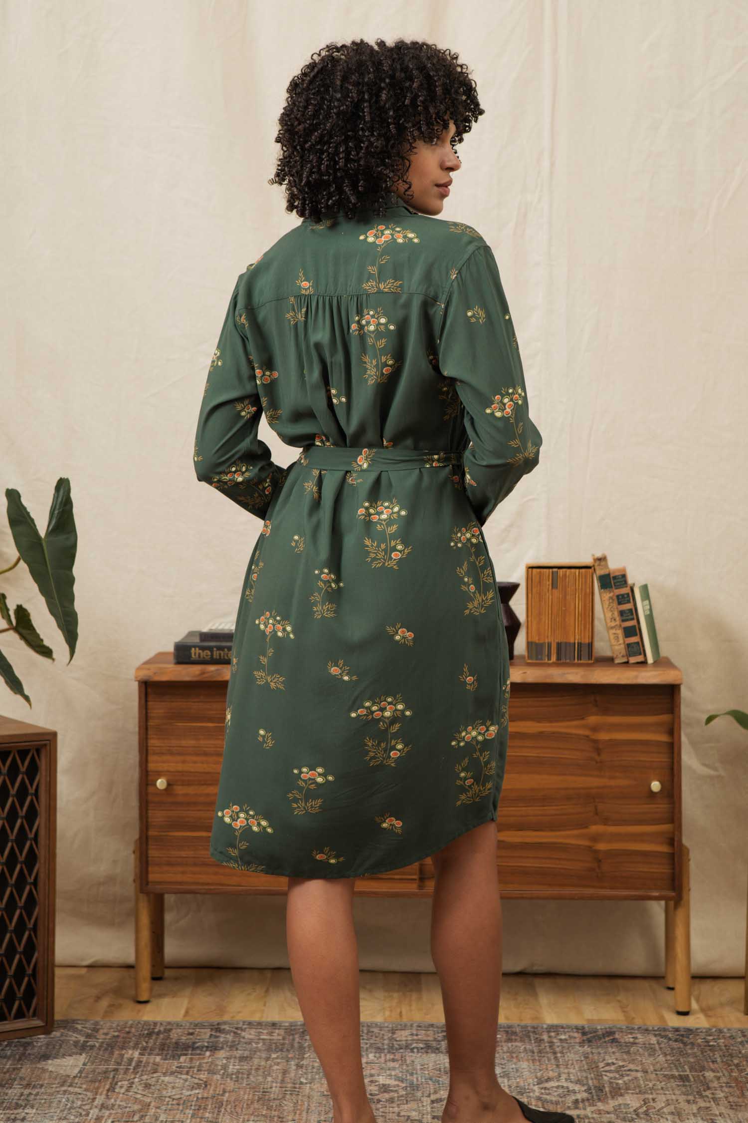 A woman with curly hair stands with her back to the camera, wearing the Bridge & Burn Emery Shirt Dress in Woodland Bloom, a green, long-sleeved, belted floral dress. She is in a cozy room with a wooden dresser, books, and plants.