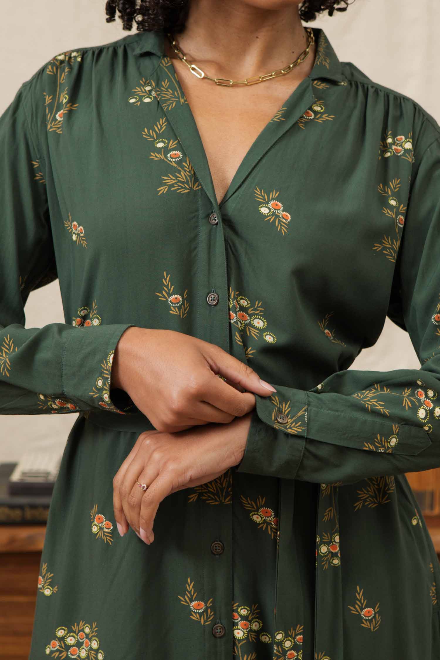 A woman in the Bridge & Burn Emery Shirt Dress in Woodland Bloom adjusts her sleeve. She accessorizes with a gold necklace and engagement ring, with her curly hair partially visible.