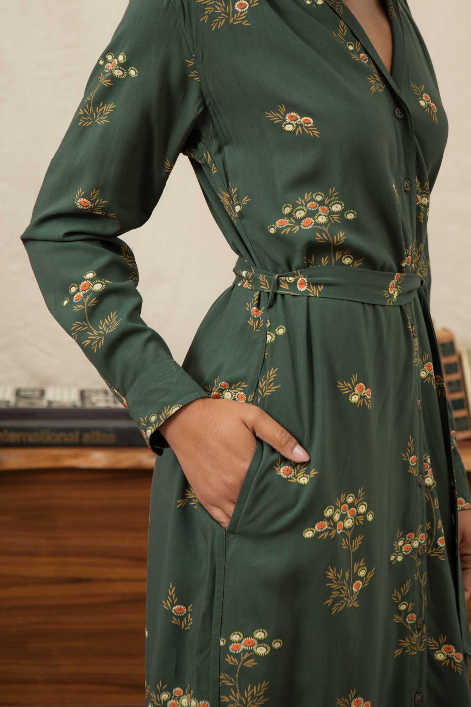 A person wears the Bridge & Burn Emery Shirt Dress in Woodland Bloom—a dark green, long-sleeved v-neck floral dress—with one hand in the pocket, standing near a wooden table and a record player.