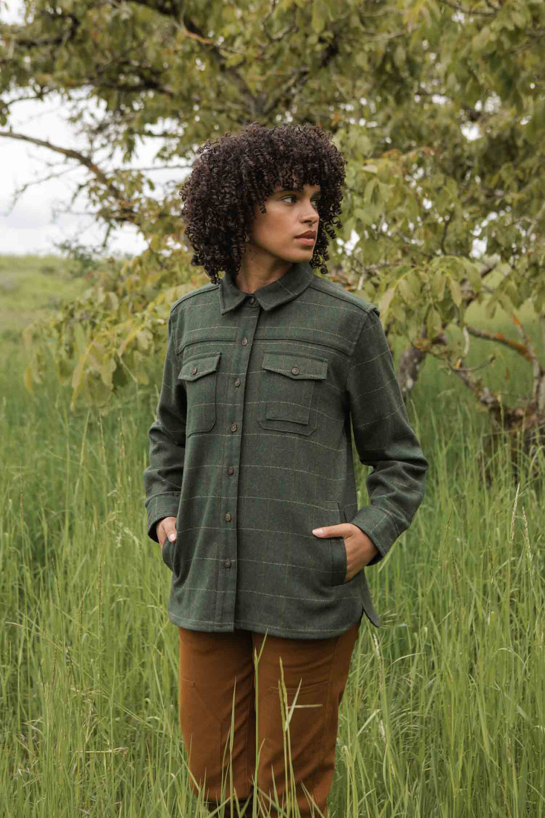 A person with curly hair stands in tall green grass, wearing the Bridge & Burn Harlow Shirt Jacket in Cypress Windowpane with brown pants, looking to the side. Leafy trees fill the background.