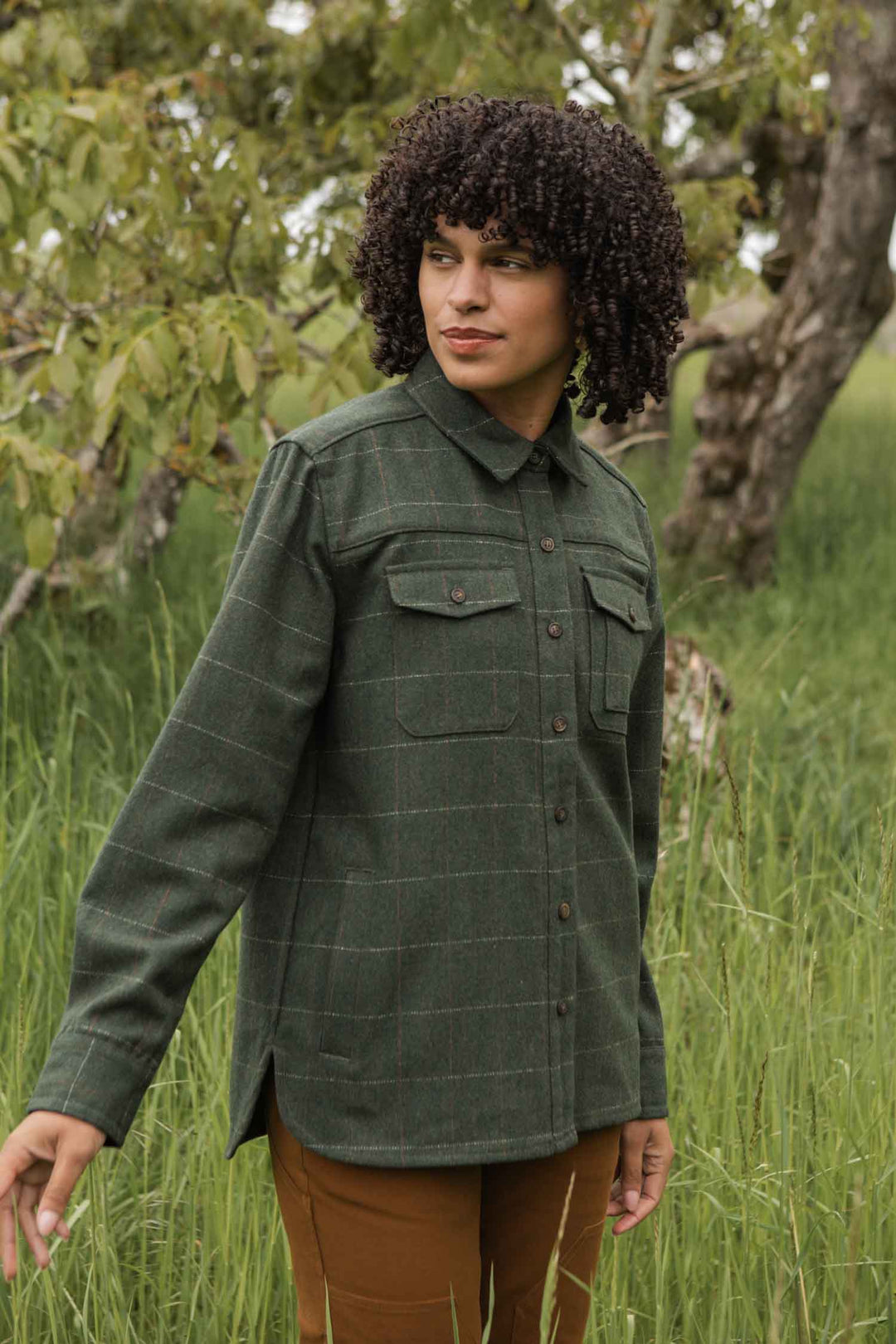 A person with curly hair stands in tall grass wearing the Bridge & Burn Harlow Shirt Jacket in Cypress Windowpane and brown pants, looking off to the side with trees and green leaves visible in the background.