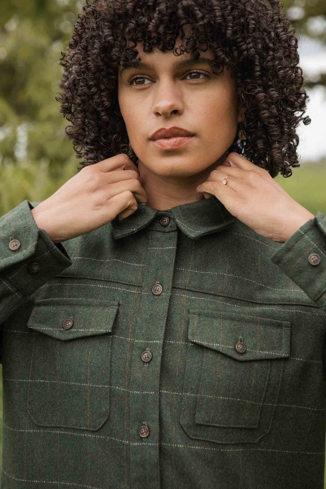 A person with curly hair adjusts the collar of the Bridge & Burn Harlow Shirt Jacket in Cypress Windowpane, a green plaid heavyweight flannel, while standing outdoors with blurred greenery in the background.