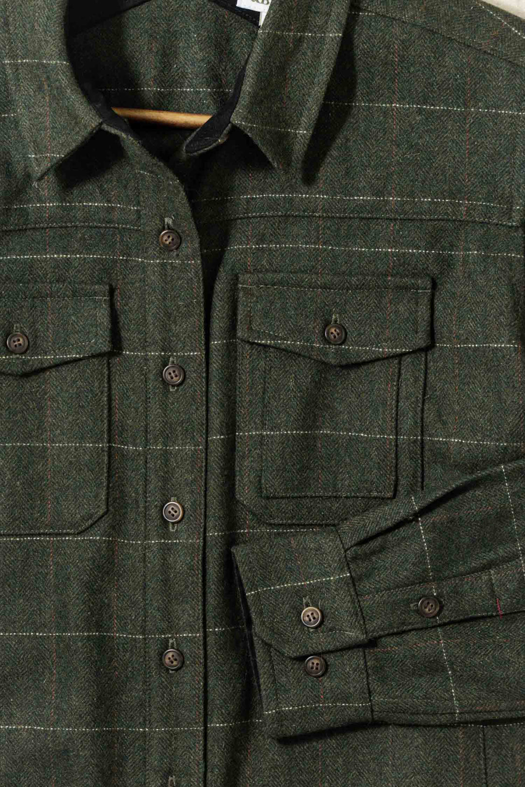 A close-up of the Bridge & Burn Harlow Shirt Jacket in Cypress Windowpane—a green plaid heavyweight flannel with two chest pockets, brown buttons, and long sleeves—displayed on a hanger.
