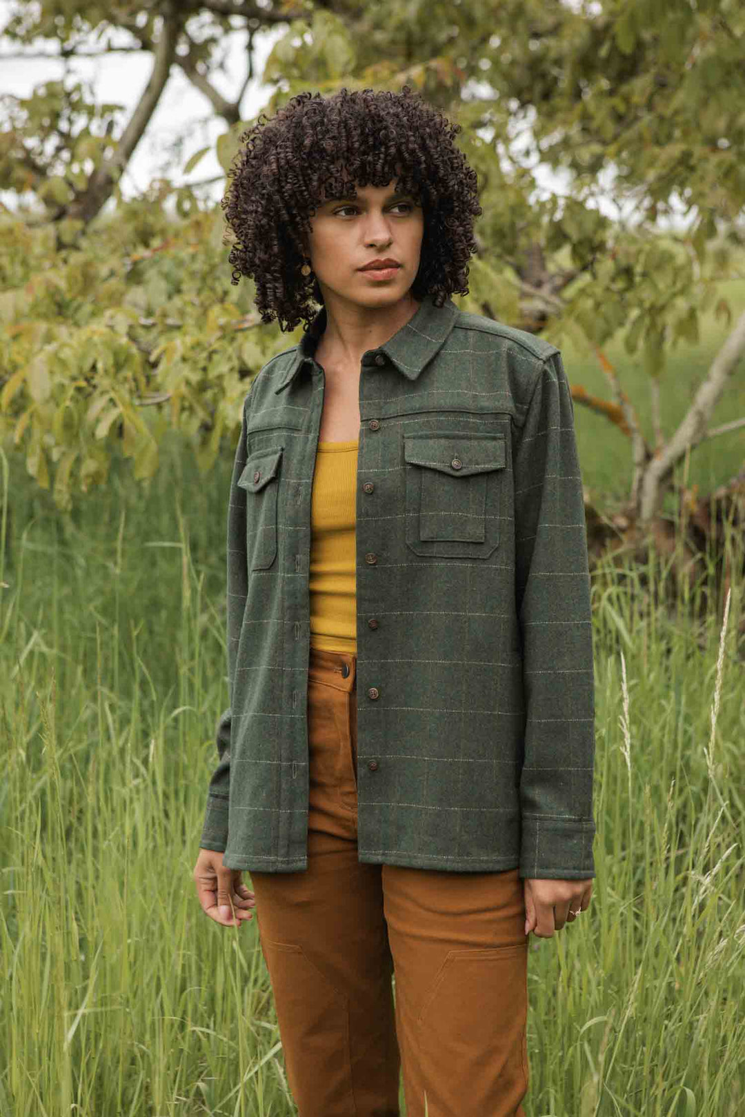 Wearing the Bridge & Burn Harlow Shirt Jacket in Cypress Windowpane, a person with curly hair pairs it with a mustard yellow top and brown pants, standing in tall green grass with trees behind.