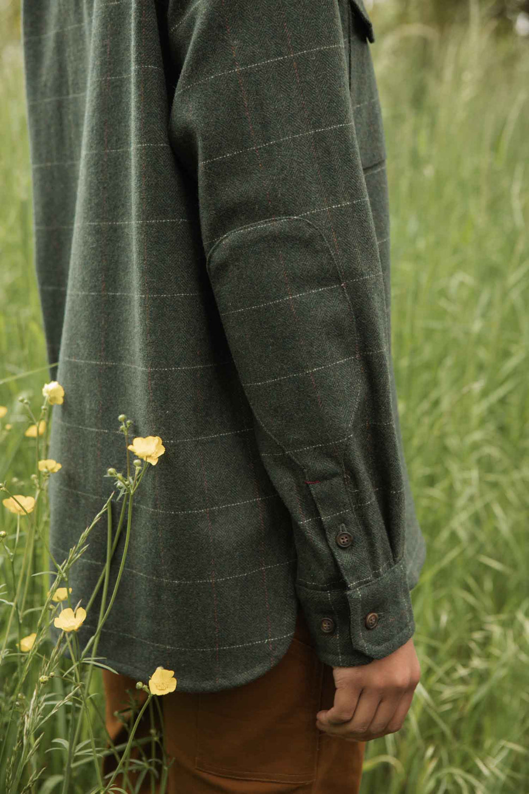 A person stands in tall grass and yellow wildflowers, wearing the Bridge & Burn Harlow Shirt Jacket in Cypress Windowpane with sleeves buttoned at the wrist. The photo focuses on the torso and arm.
