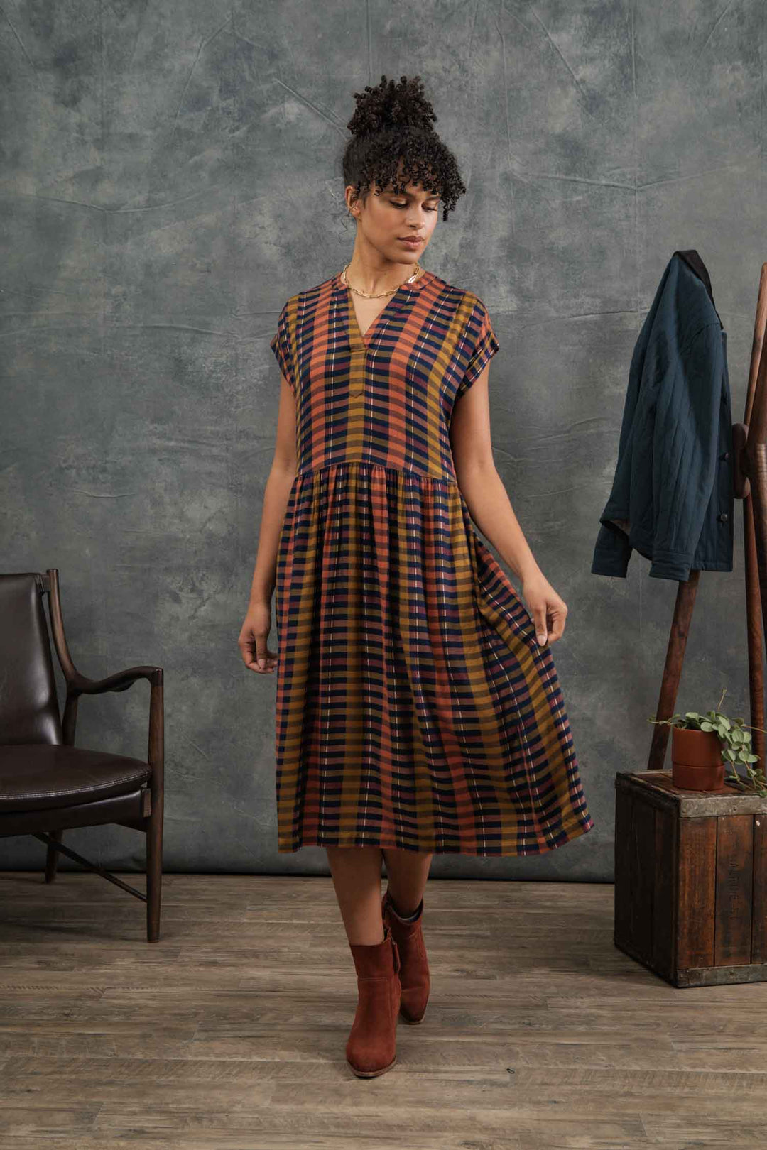 A woman stands indoors on a wooden floor wearing the Bridge & Burn Ira Dress in Alpenglow Stripe with rust ankle boots; a green jacket and potted plant are visible in the background.
