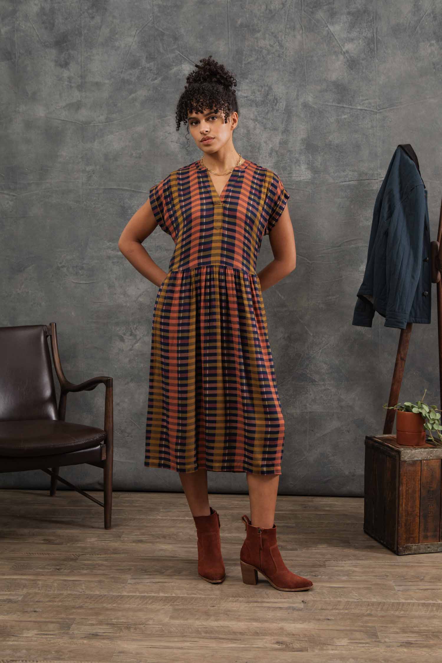 The Ira Dress by Bridge & Burn features knee-length, short dolman sleeves, a v-neck, Alpenglow Stripe pattern in mustard yellow, rust orange, navy blue, and black. Made from eco-friendly viscose with a gathered waist and relaxed fit.