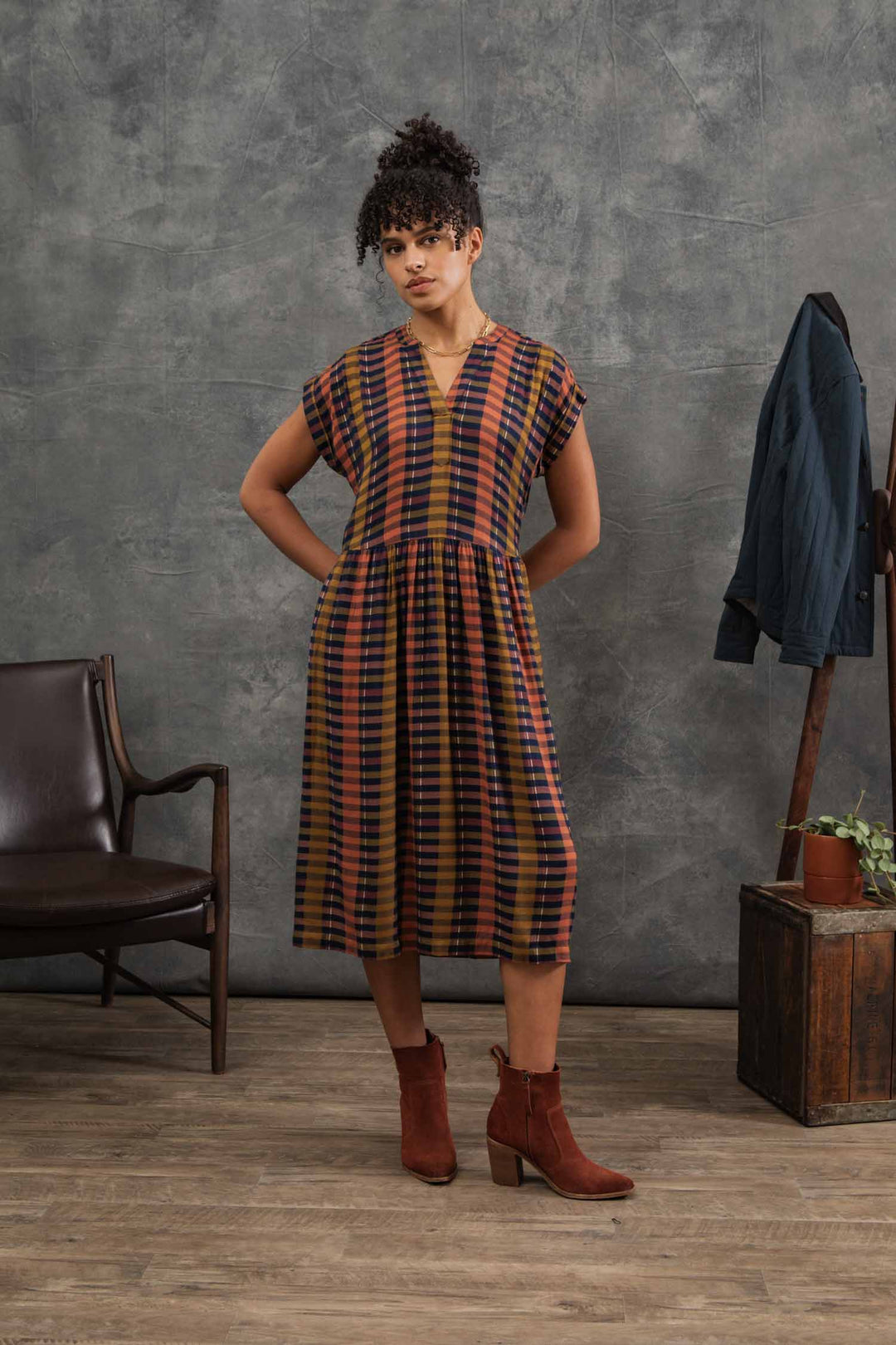 A woman stands indoors wearing the Bridge & Burn Ira Dress in Alpenglow Stripe with brown ankle boots. Behind her, a jacket hangs on a wooden crate next to a plant, and a leather chair sits against a gray textured wall.
