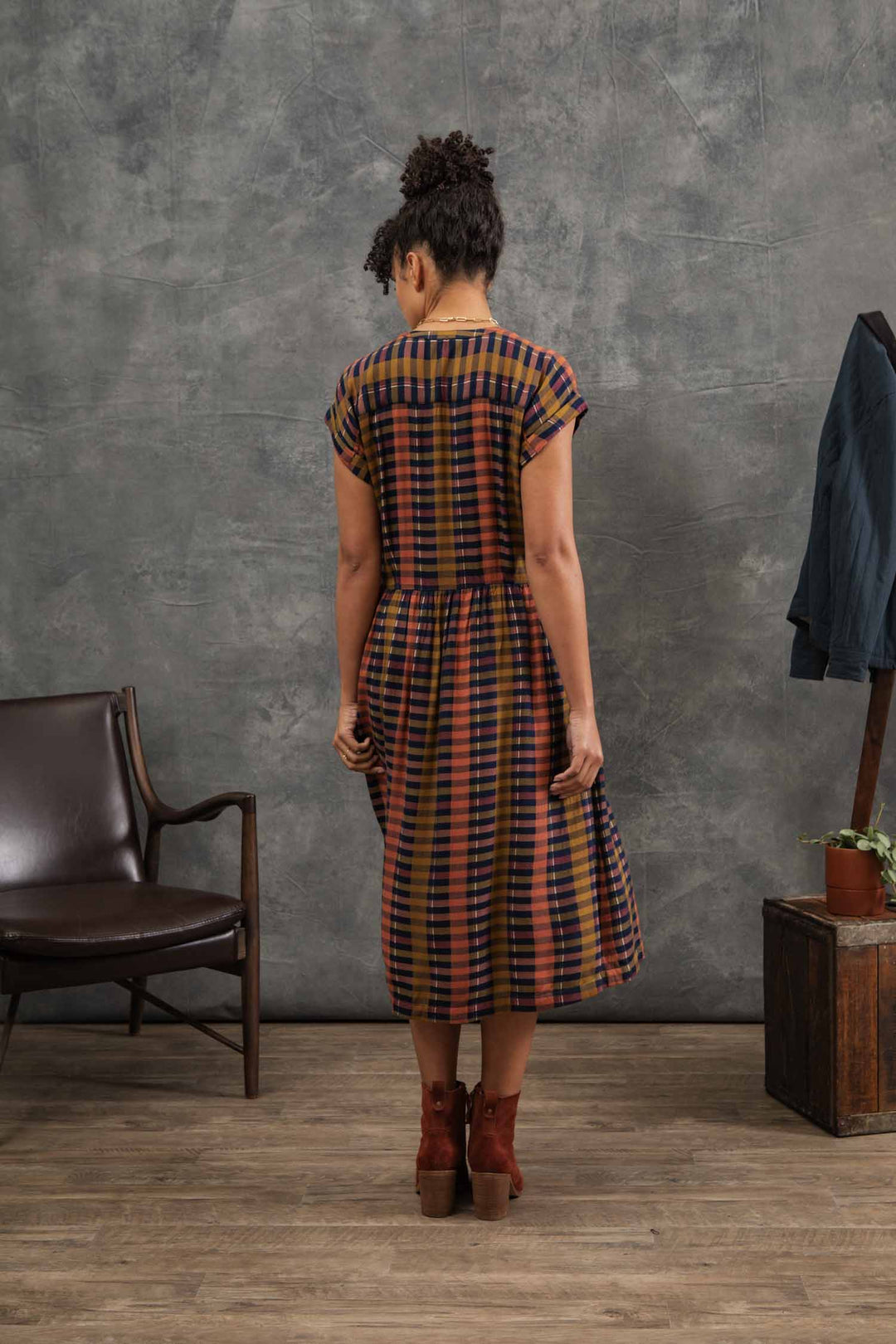 A woman stands indoors with her back to the camera, wearing the Bridge & Burn Ira Dress in Alpenglow Stripe—a colorful eco-friendly viscose pullover—paired with brown ankle boots. Nearby are a chair, jacket, and potted plant.