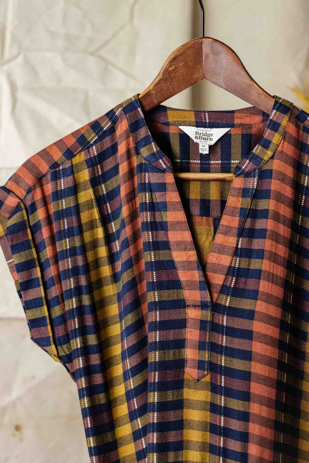 The Bridge & Burn Ira Dress in Alpenglow Stripe, a short-sleeved pullover with dolman sleeves and a plaid pattern in navy, mustard, and rust, hangs on a wooden hanger against a light textured background.