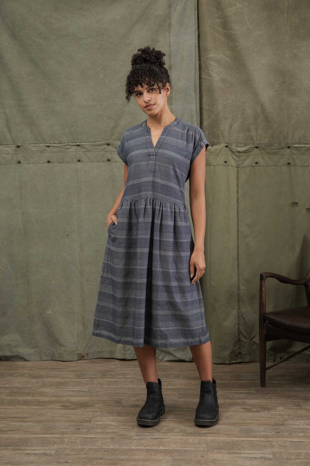 A woman stands indoors wearing the Bridge & Burn Ira Dress in Cascade Weave, a short-sleeved, knee-length organic cotton pullover with pockets and gray stripes. She pairs it with black ankle boots beside a wooden chair and green fabric backdrop.