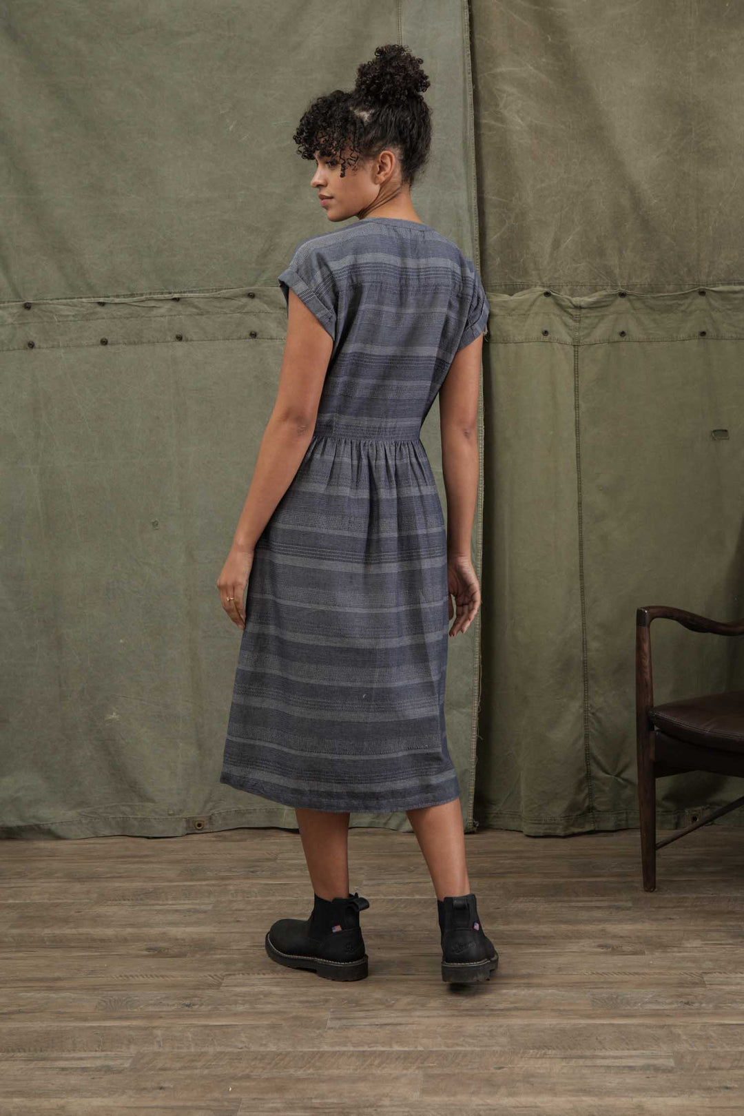 A woman stands indoors on a wooden floor, facing away. She wears the Bridge & Burn Ira Dress in Cascade Weave with black ankle boots. A green curtain hangs in the background beside a brown chair.