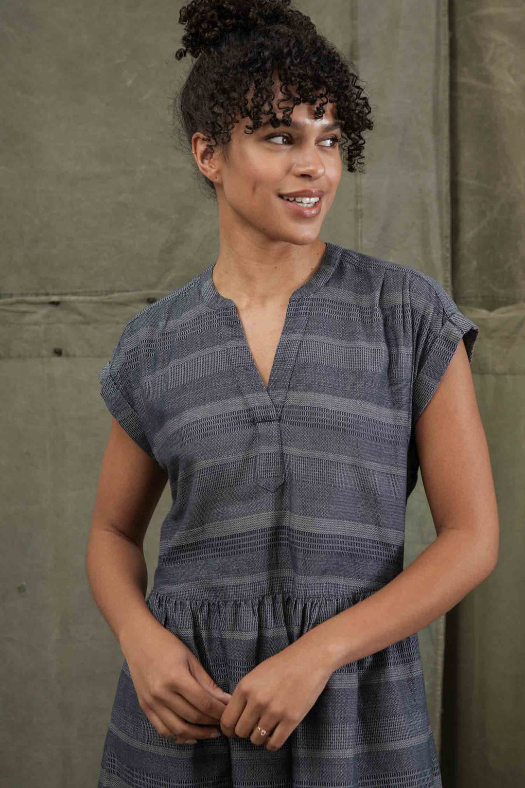 A woman with curly hair in an updo wears the Bridge & Burn Ira Dress in Cascade Weave. The short-sleeved, gray striped pullover features a V-neck. She stands before a plain olive green backdrop, smiling and looking to her left.