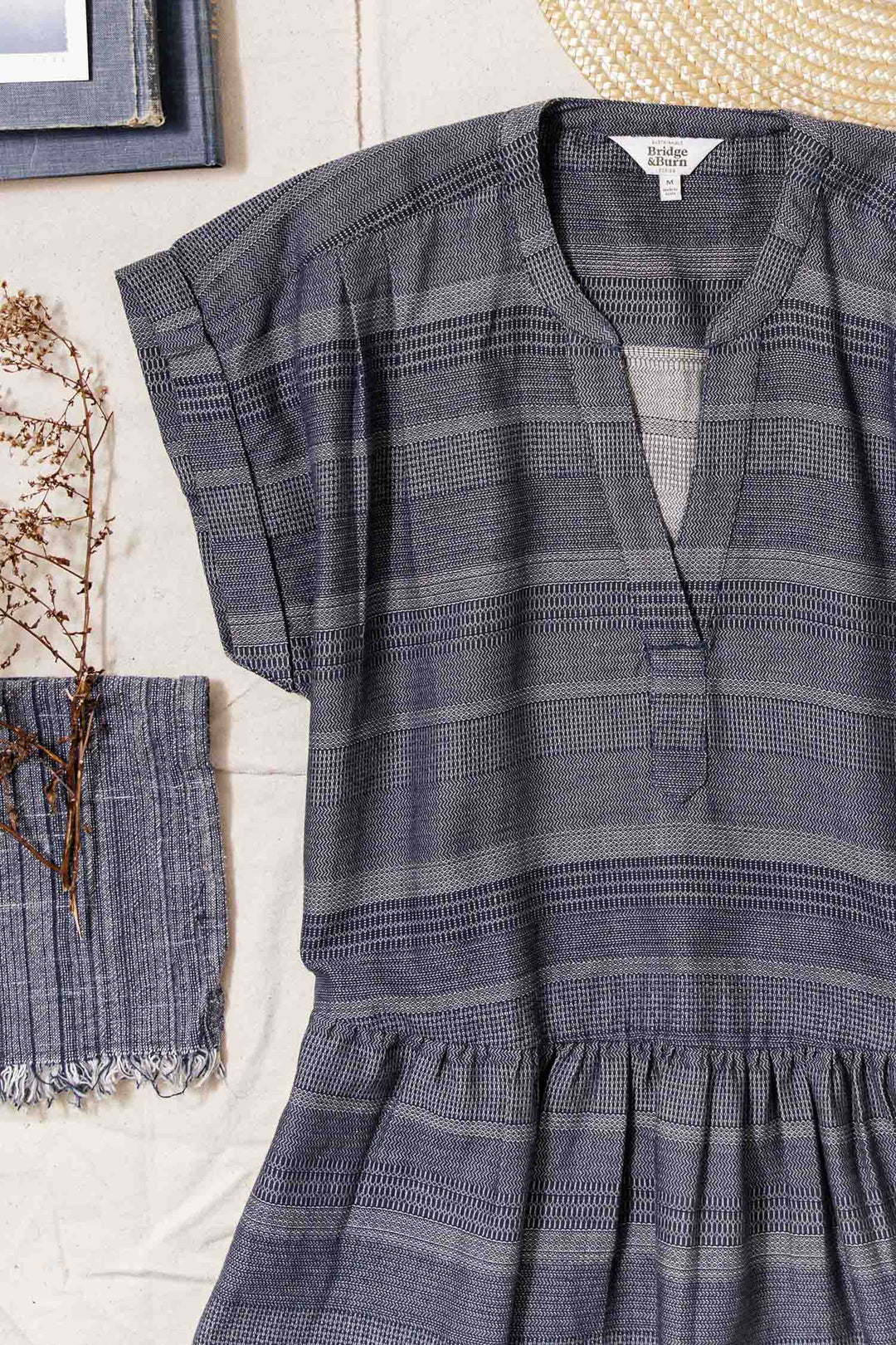The Bridge & Burn Ira Dress / Cascade Weave, a short-sleeved blue-gray organic cotton dress with horizontal textured stripes, is laid flat on a beige surface beside a fringed cloth, dried branches, straw hat, and part of a blue-framed picture.