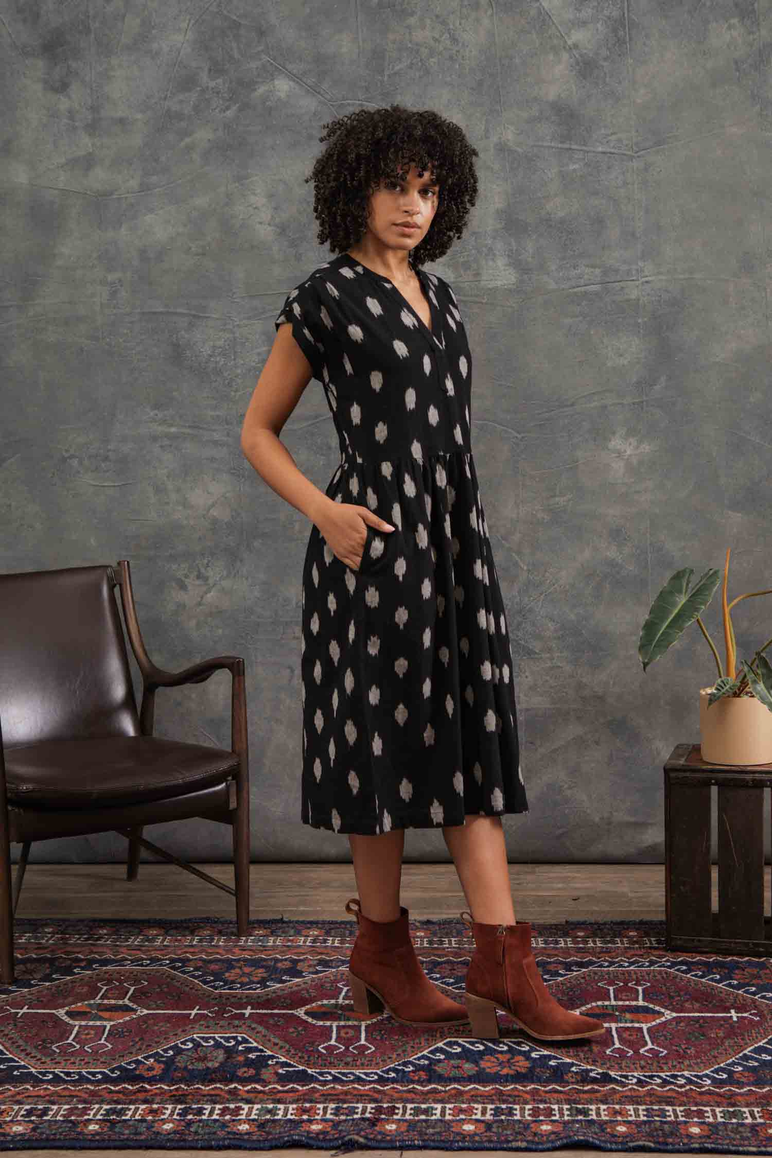 A woman with curly hair models the Bridge & Burn Ira Dress in Eclipse Ikat—a black 100% cotton pullover with dolman sleeves and a white dotted pattern—standing by a brown chair and plant on a patterned rug before a gray backdrop.