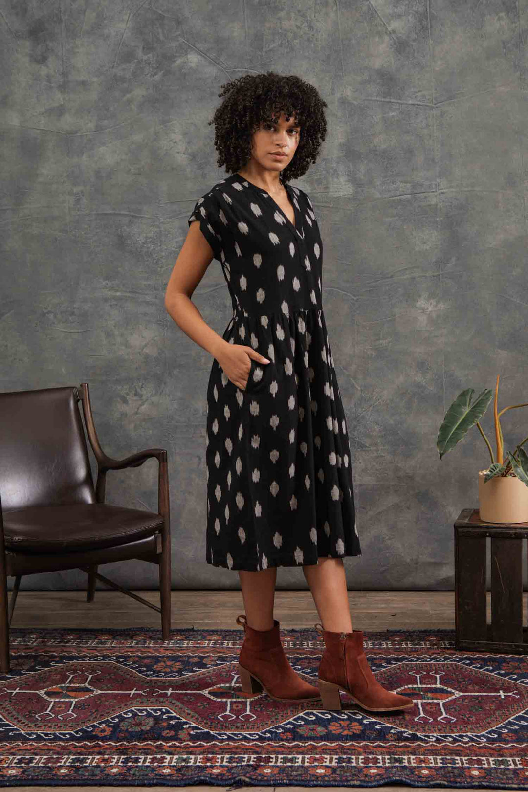 A woman with curly hair models the Bridge & Burn Ira Dress in Eclipse Ikat—a black 100% cotton pullover with dolman sleeves and a white dotted pattern—standing by a brown chair and plant on a patterned rug before a gray backdrop.