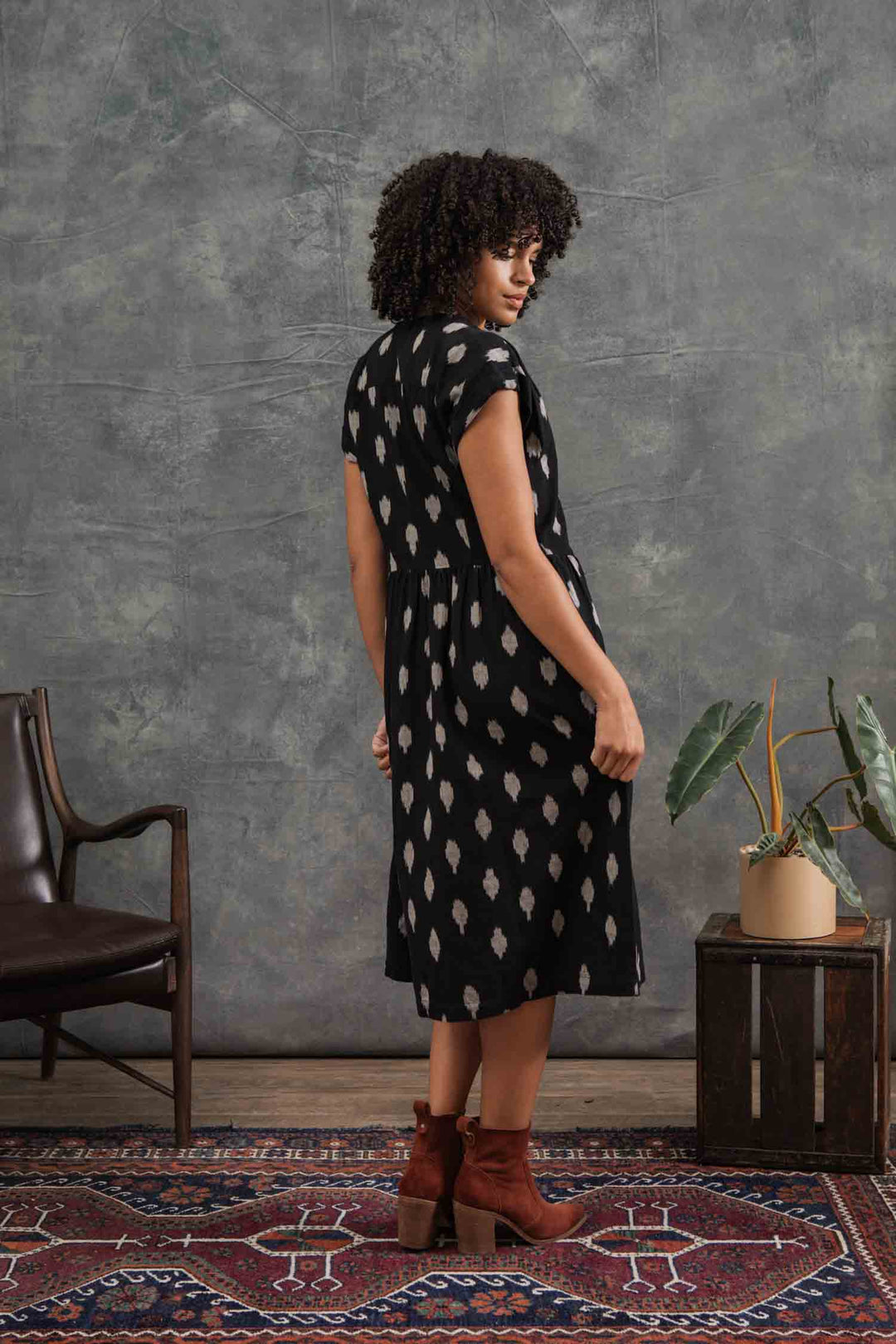A woman with curly hair stands sideways, wearing the Bridge & Burn Ira Dress in Eclipse Ikat with dolman sleeves and brown ankle boots. She is on a patterned rug beside a dark chair and potted plant, against a gray textured wall.